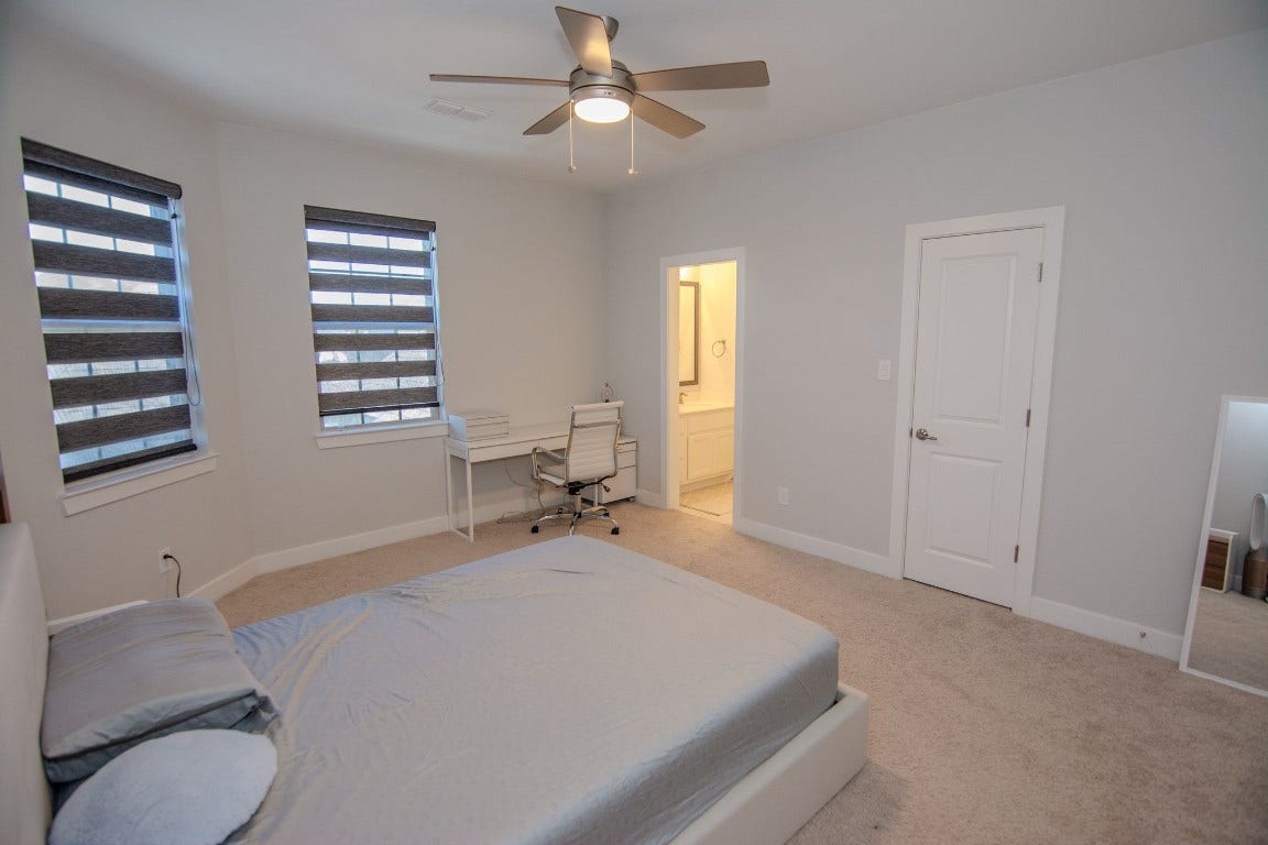5423 Priamus Drive, Missouri City, TX 77459 This photo showcases a spacious, modern bedroom with neutral walls and carpet. It features two windows with stylish blinds, a ceiling fan, and a small workspace. There's an en-suite bathroom and a closet, providing convenience and comfort.
