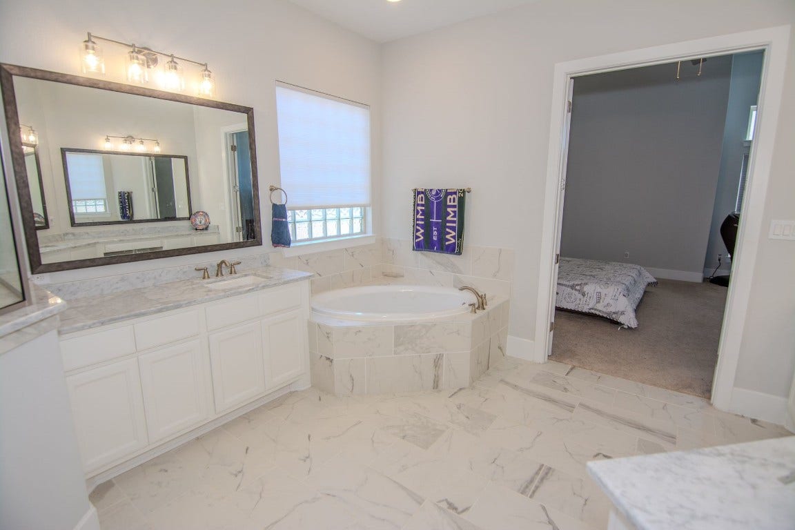 5423 Priamus Drive, Missouri City, TX 77459 This photo showcases a spacious bathroom featuring a large mirror, elegant vanity with marble countertops, and a luxurious corner bathtub. The room is well-lit with natural light from a window, and there's an adjoining door leading to a bedroom. The design is modern with a clean and bright aesthetic.