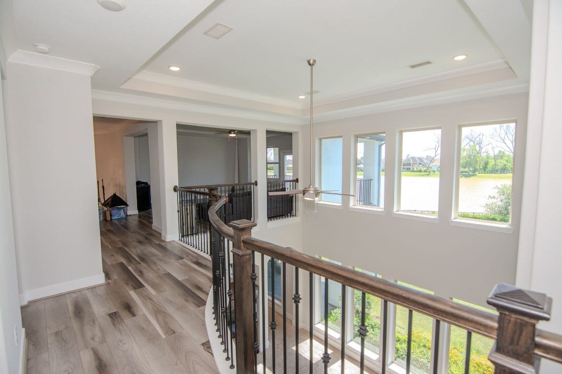 5423 Priamus Drive, Missouri City, TX 77459 This photo showcases a bright, open hallway with modern wood flooring and a stylish railing. Large windows provide natural light and offer a scenic view of a lake, creating a serene atmosphere.