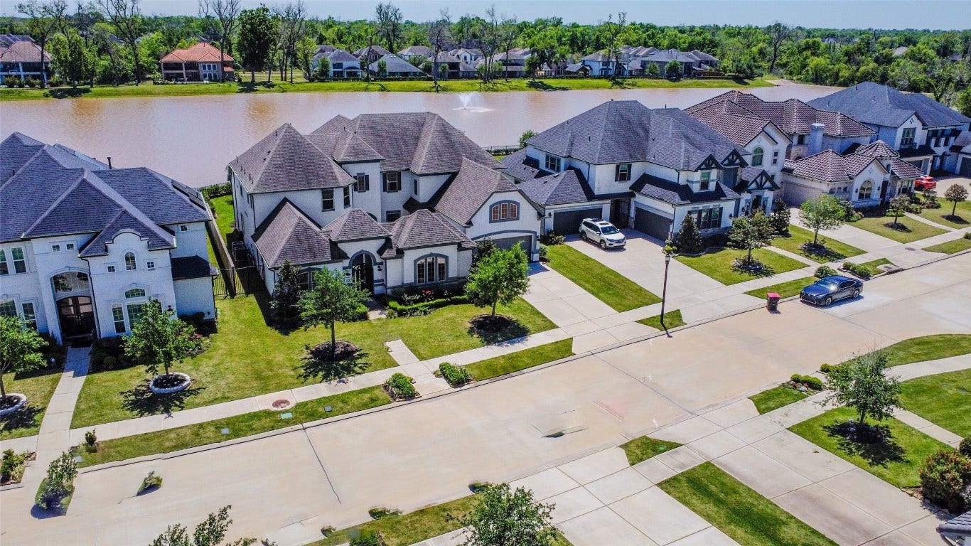 5423 Priamus Drive, Missouri City, TX 77459 This photo showcases a serene residential neighborhood with elegant, large homes lining a tree-dotted street. The houses have well-maintained lawns and are situated near a picturesque lake, offering scenic water views and a peaceful atmosphere.