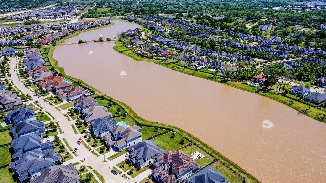 5423 Priamus Drive, Missouri City, TX 77459 This aerial photo showcases a picturesque residential neighborhood with houses lining a serene, brownish lake featuring fountains. The area is lush with greenery and offers a tranquil suburban setting, ideal for families seeking a peaceful environment.