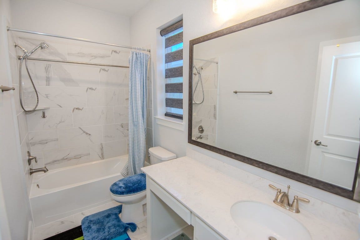 5423 Priamus Drive, Missouri City, TX 77459 This bathroom features a clean design with a tiled shower and tub combo, a large framed mirror above a marble countertop, and modern fixtures. A window allows natural light, and the space includes a blue-themed bath mat and shower curtain.