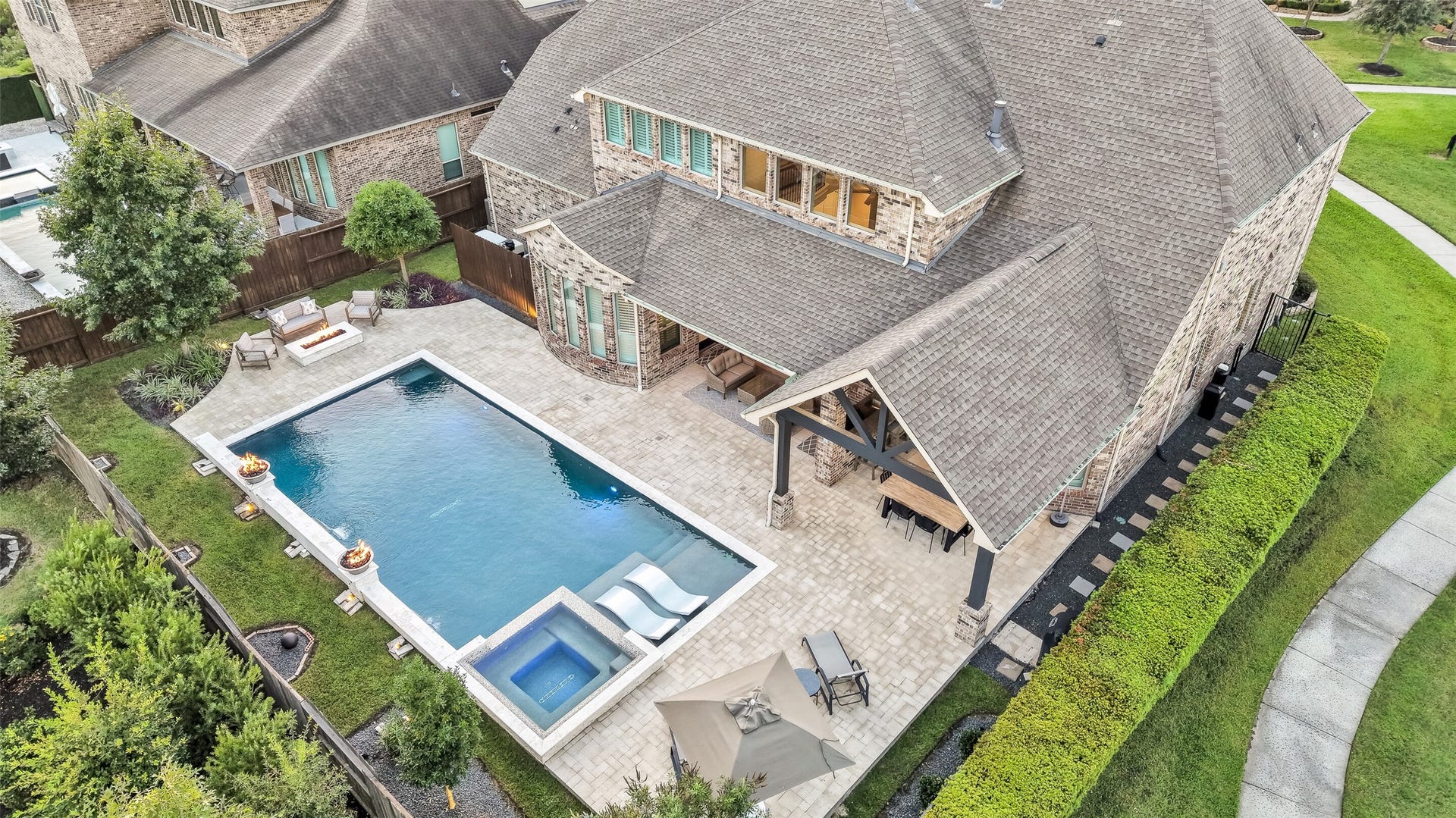 This aerial view captures the full scope of the private backyard retreat: a custom heated pool and raised spa, a spacious paver patio, built-in fire bowls, fire pit area and covered outdoor living and dining areas all ready for effortless indoor-outdoor entertaining.