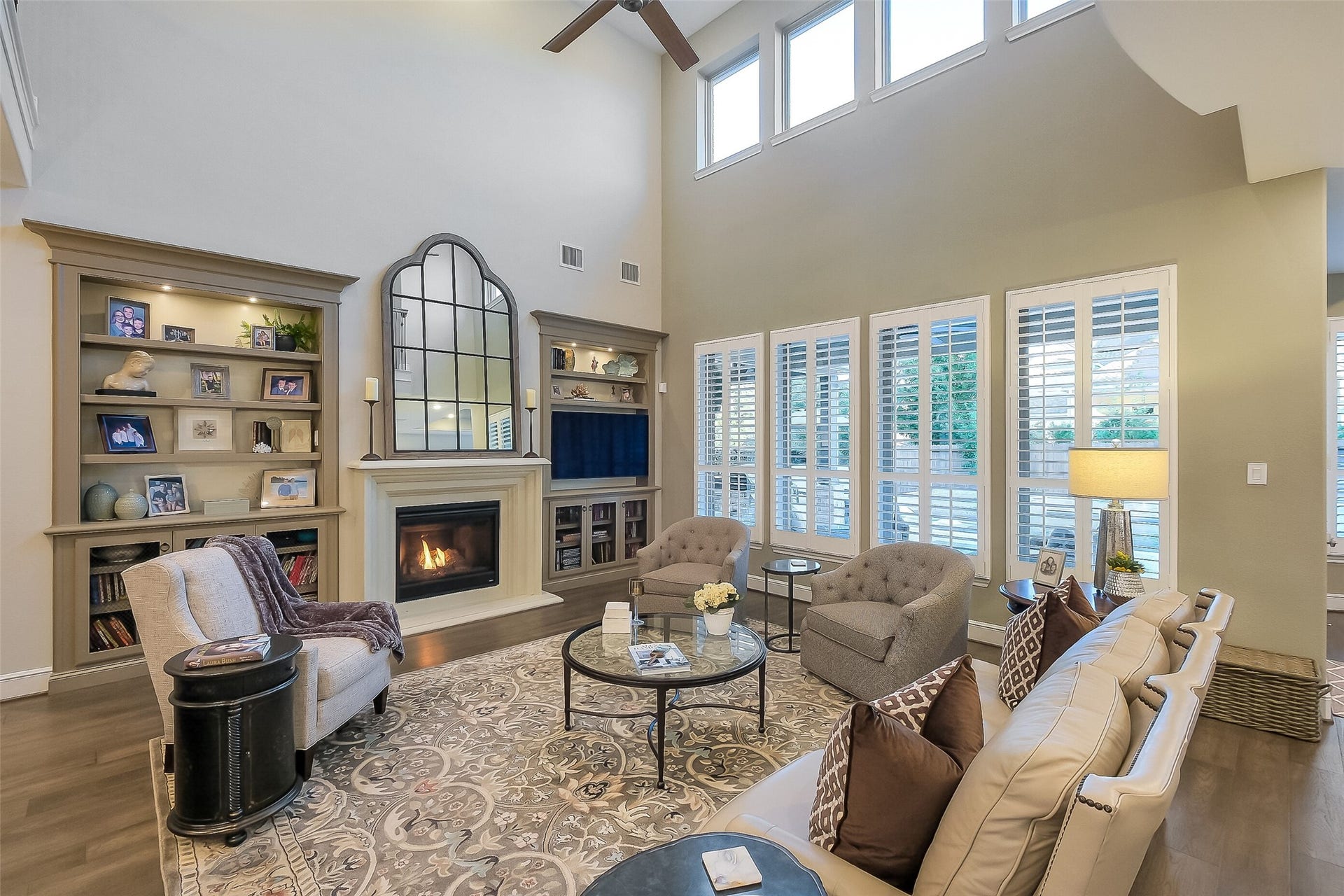 Soaring two-story ceilings and a wall of plantation-shuttered windows fill this living room with natural light. A cozy gas fireplace and custom built-in shelving create a warm, welcoming gathering space.