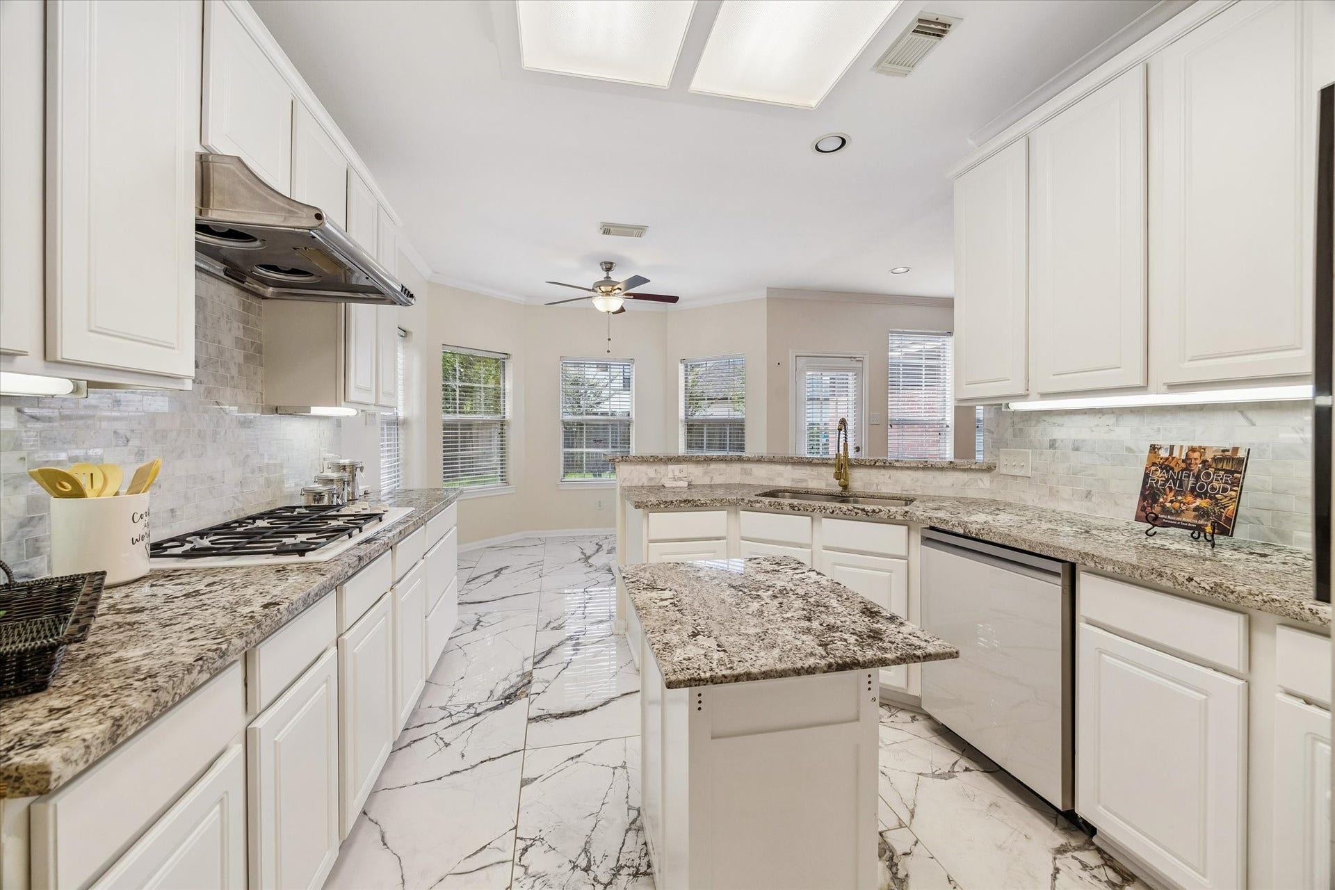 1115 Mulberry Lane, Bellaire, TX 77401 Eelegant granite countertops, sleek white cabinetry, and modern appliances, complemented by a bright, airy breakfast nook with large windows and a ceiling fan.