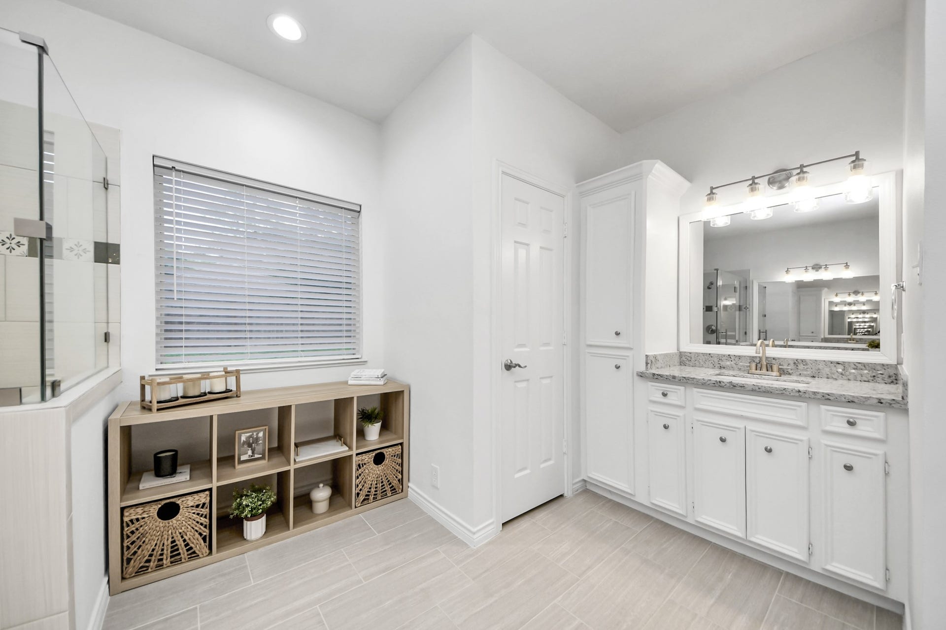 Elegant bathroom with a spacious vanity featuring granite countertops, ample cabinetry, and modern lighting. A stylish shelving unit and large window enhance the airy ambiance.