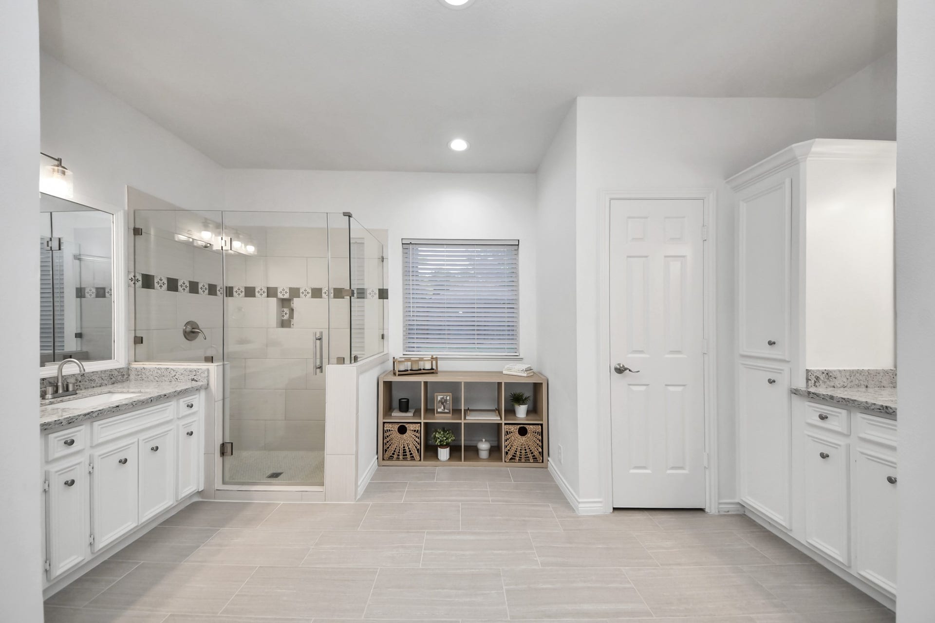 Separate vanities on opposite walls give each person their own space in this primary bath, finished with granite countertops and crisp white cabinetry. The frameless glass walk-in shower features a decorative accent tile band and a built-in niche. Large-format tile flooring, a tall linen cabinet, and natural light round out the room.