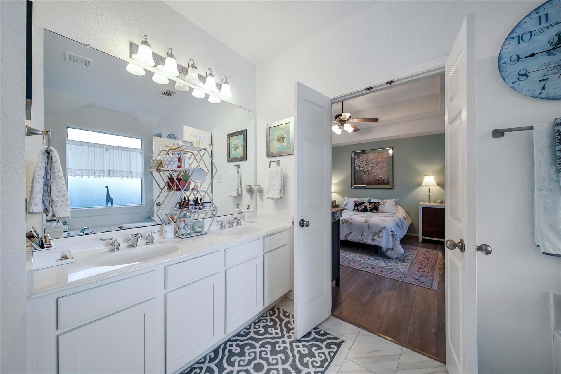 This is a spacious and well-lit master bedroom featuring a large bed, hardwood floors, a ceiling fan, and tasteful decor. The room includes a cozy sitting area and ample natural light from a broad window with curtains.
