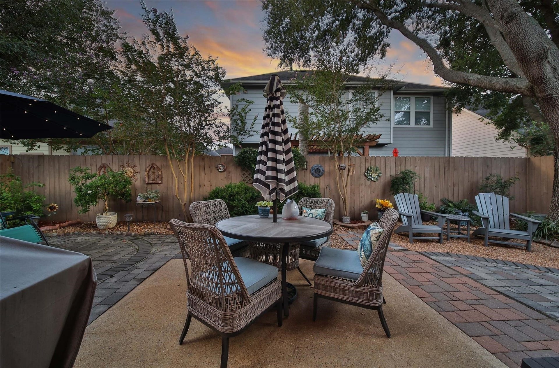 This is a cozy backyard with a brick patio and comfortable seating under the shade of a large tree, enclosed by a privacy fence, adjacent to a house with a warm exterior.