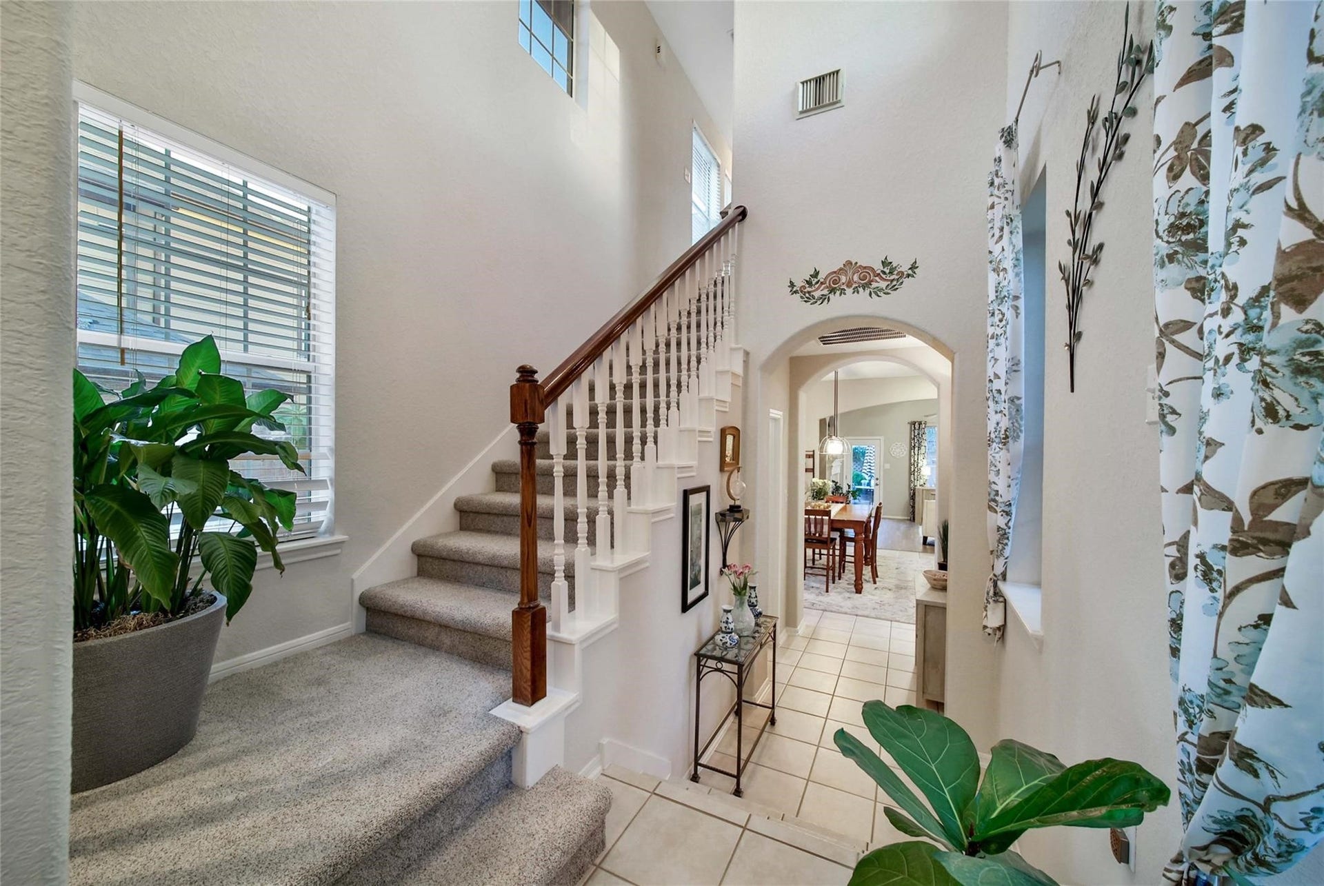 This is a bright, inviting entryway featuring a staircase with wooden banisters, high ceilings, large windows, and an open archway leading to other rooms. The space has a welcoming atmosphere.