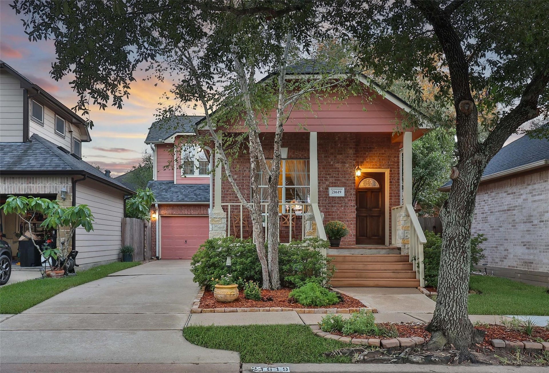 This is a charming two-story brick home with an attached garage, featuring a landscaped front yard and a welcoming porch, captured during a beautiful sunset.