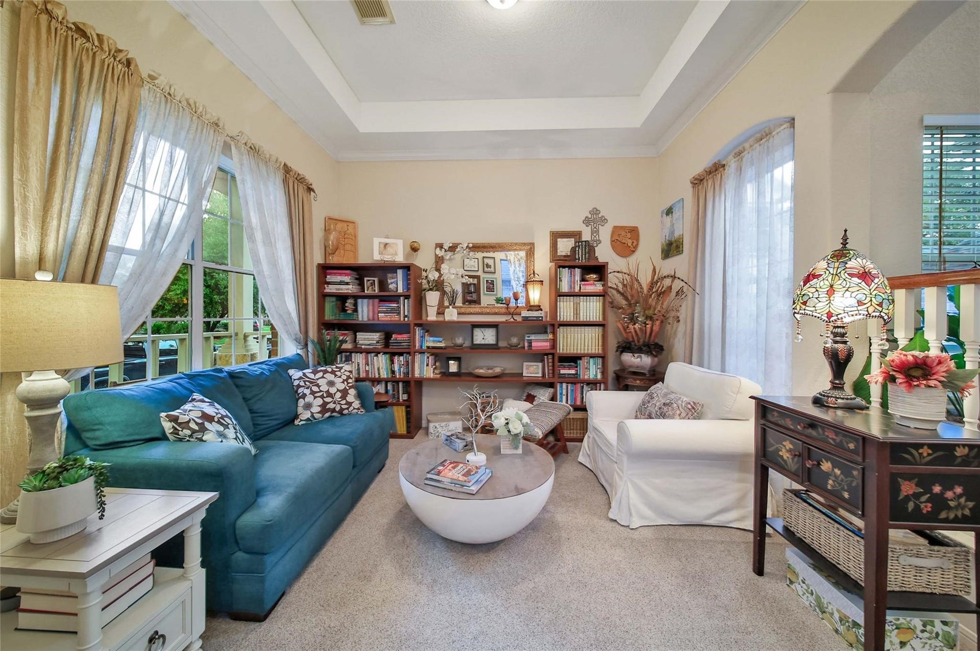 This is a cozy, well-lit living room featuring a high ceiling, a large window with flowing curtains