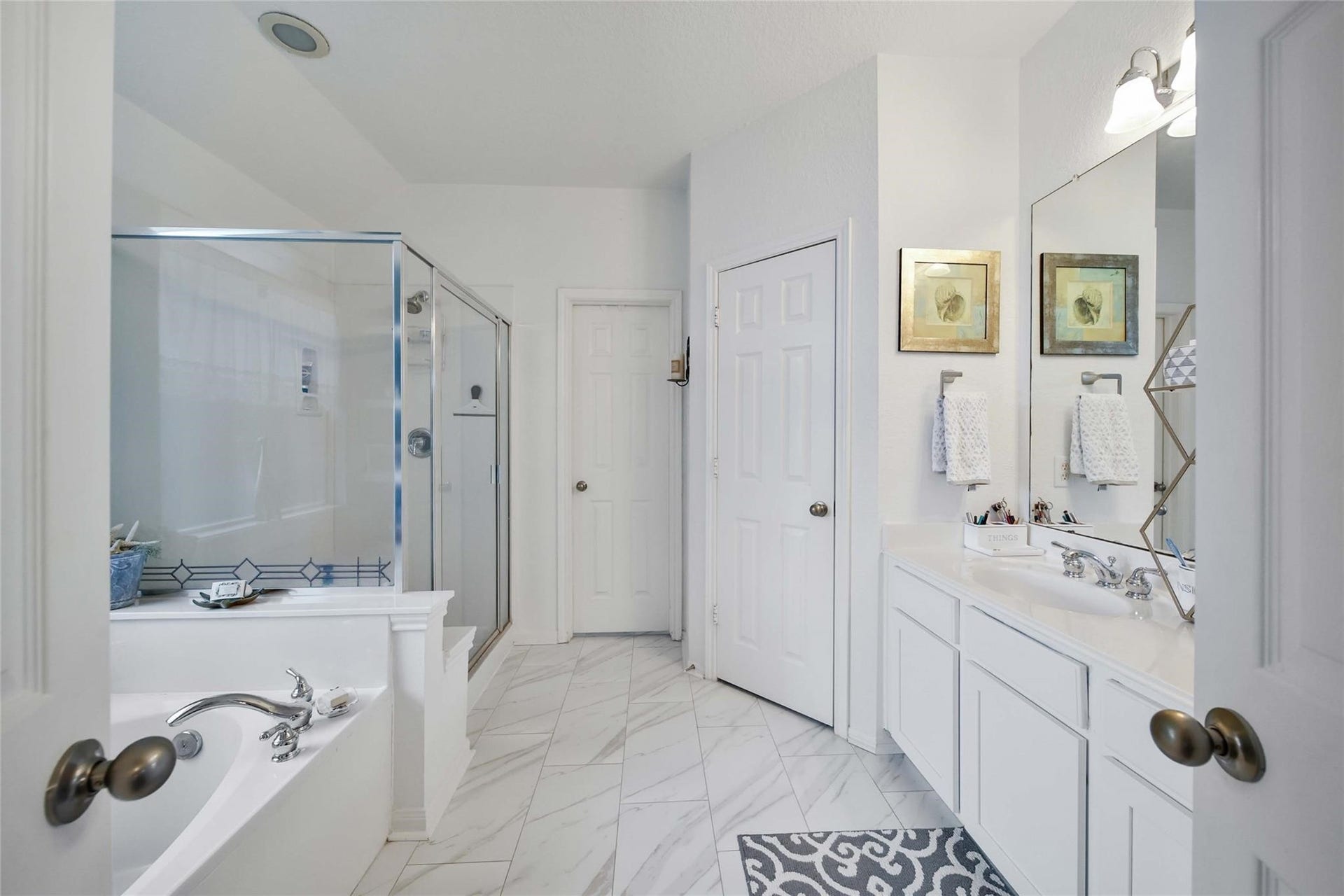 This is a bright, clean primary bathroom featuring a large vanity with dual sinks, ample storage space, and a full-width mirror with overhead lighting. The room has a tiled floor and offers both a shower and a separate tub.