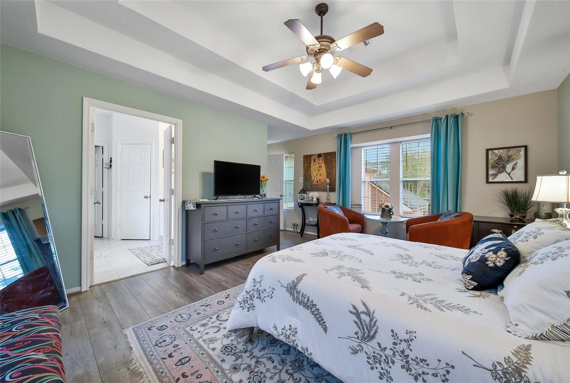 This is a well-lit hallway featuring a large window with horizontal blinds, neutral carpeting, and light-colored walls adorned with artwork, leading towards additional rooms.