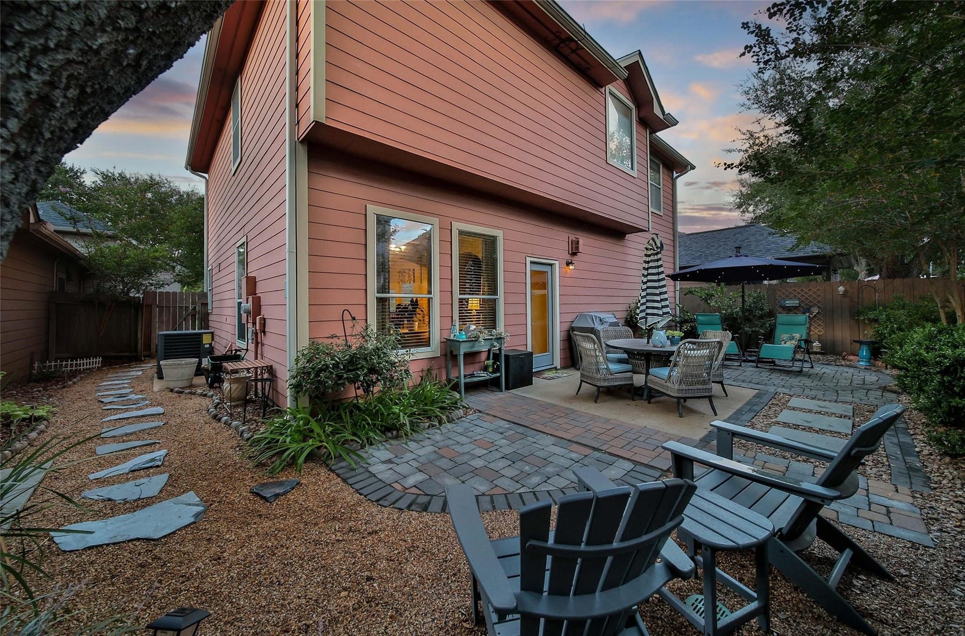 cozy backyard patio with a circular brick layout, complemented by two comfortable reclining chairs under a large umbrella. The area is surrounded by a wooden fence, offering privacy, and features lush greenery that adds to the tranquil atmosphere, ideal for relaxation or entertaining.