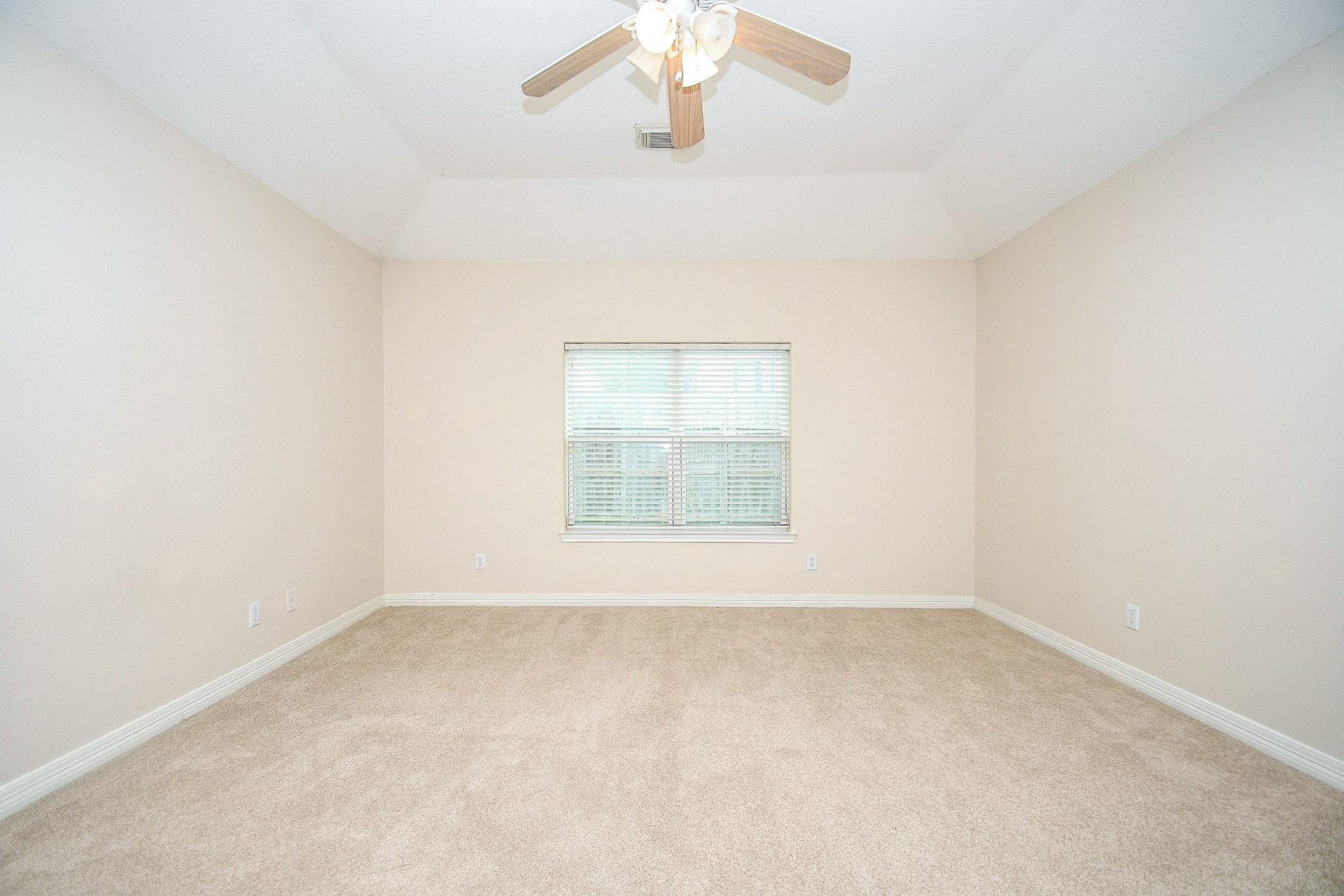 This bright, spacious room features neutral walls, plush carpet, a central window with blinds, and a ceiling fan, offering a cozy and versatile space.