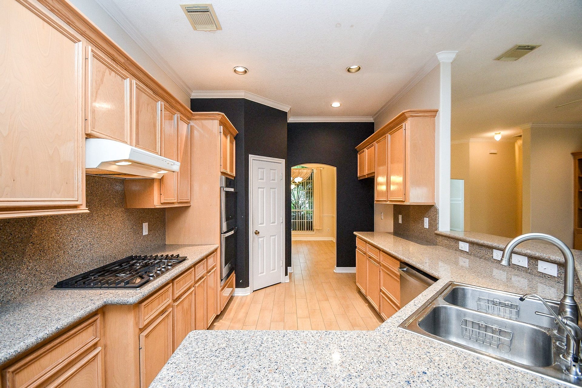 This kitchen features light wood cabinetry, granite countertops, and a built-in gas stove. It has ample storage, a double sink, and an open layout leading to a dining area. The lighting is recessed, and the flooring is hardwood.