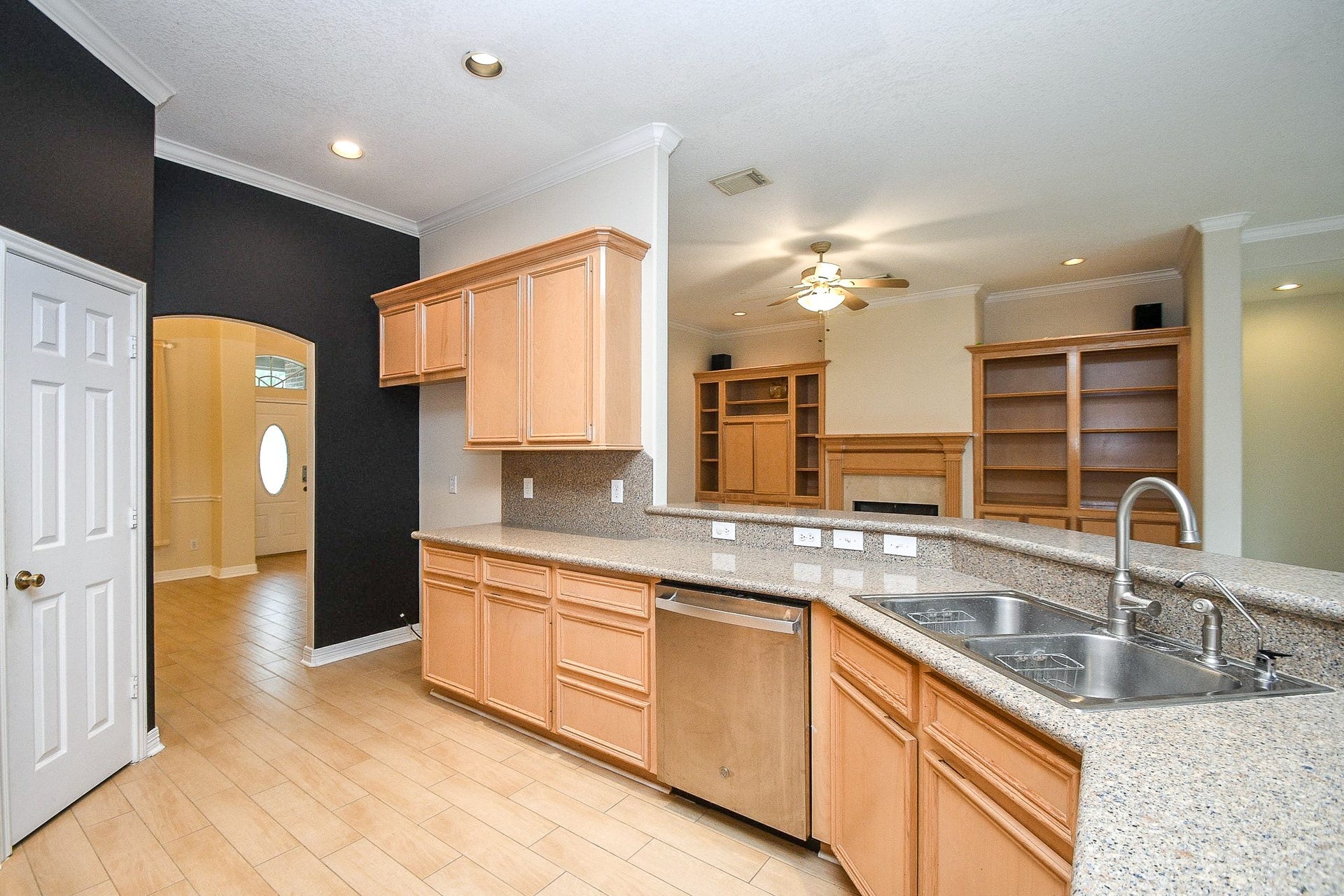 Bright and spacious kitchen with light wood cabinets, granite countertops, and a double sink. An open layout leads to a living area with built-in shelving and a fireplace. Elegant archway and ample natural light enhance the welcoming atmosphere.
