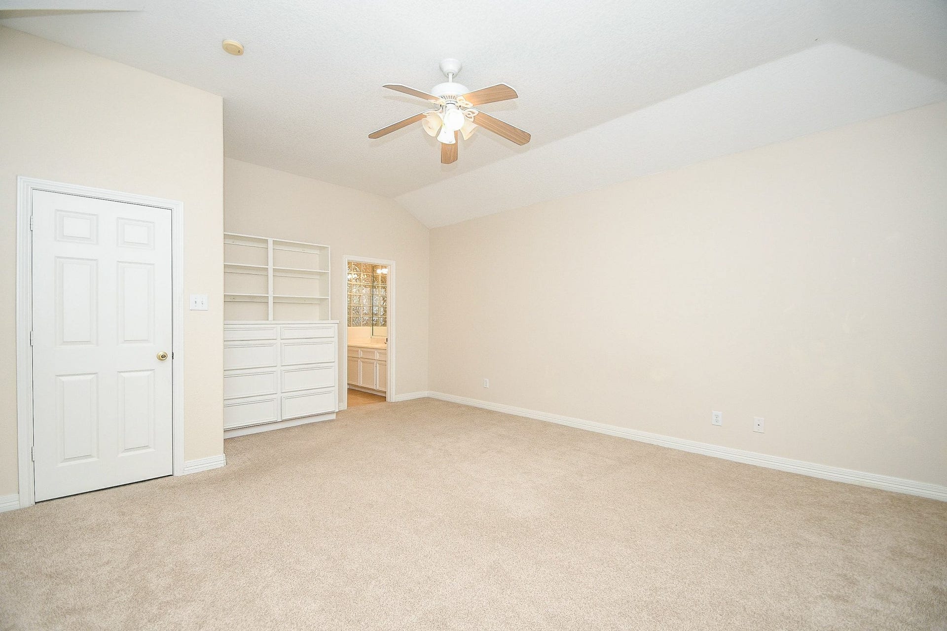 Bright, spacious room with neutral walls and carpet, featuring a ceiling fan. Includes built-in shelves and a view into a bathroom, offering convenience and ample storage.