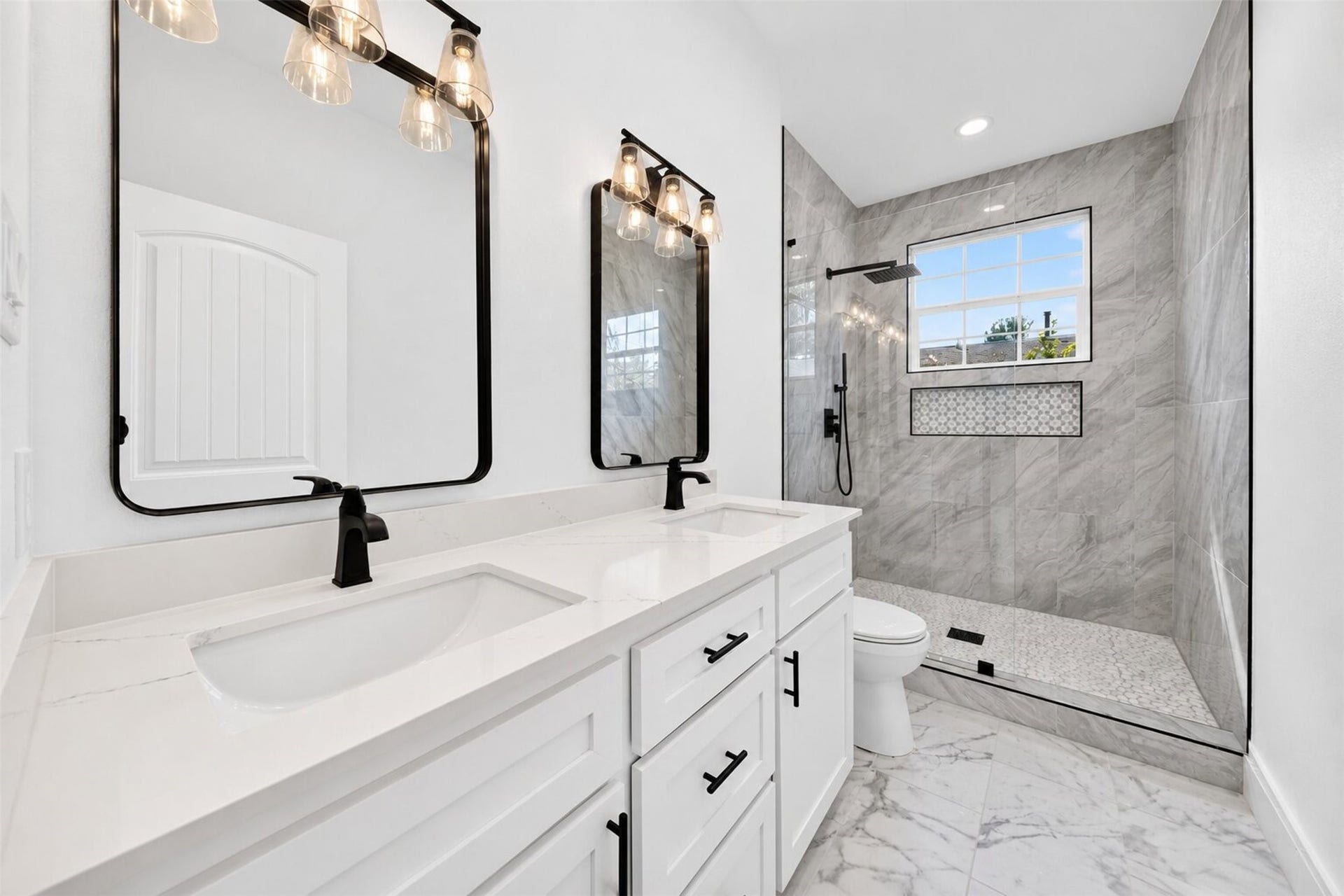 The contemporary bathroom boasts a stylish design, featuring a pristine white vanity, chic black fixtures, and a bathtub adorned with sophisticated gray tile surround. Natural light gently streams through a window, contributing to the clean and luminous ambiance.