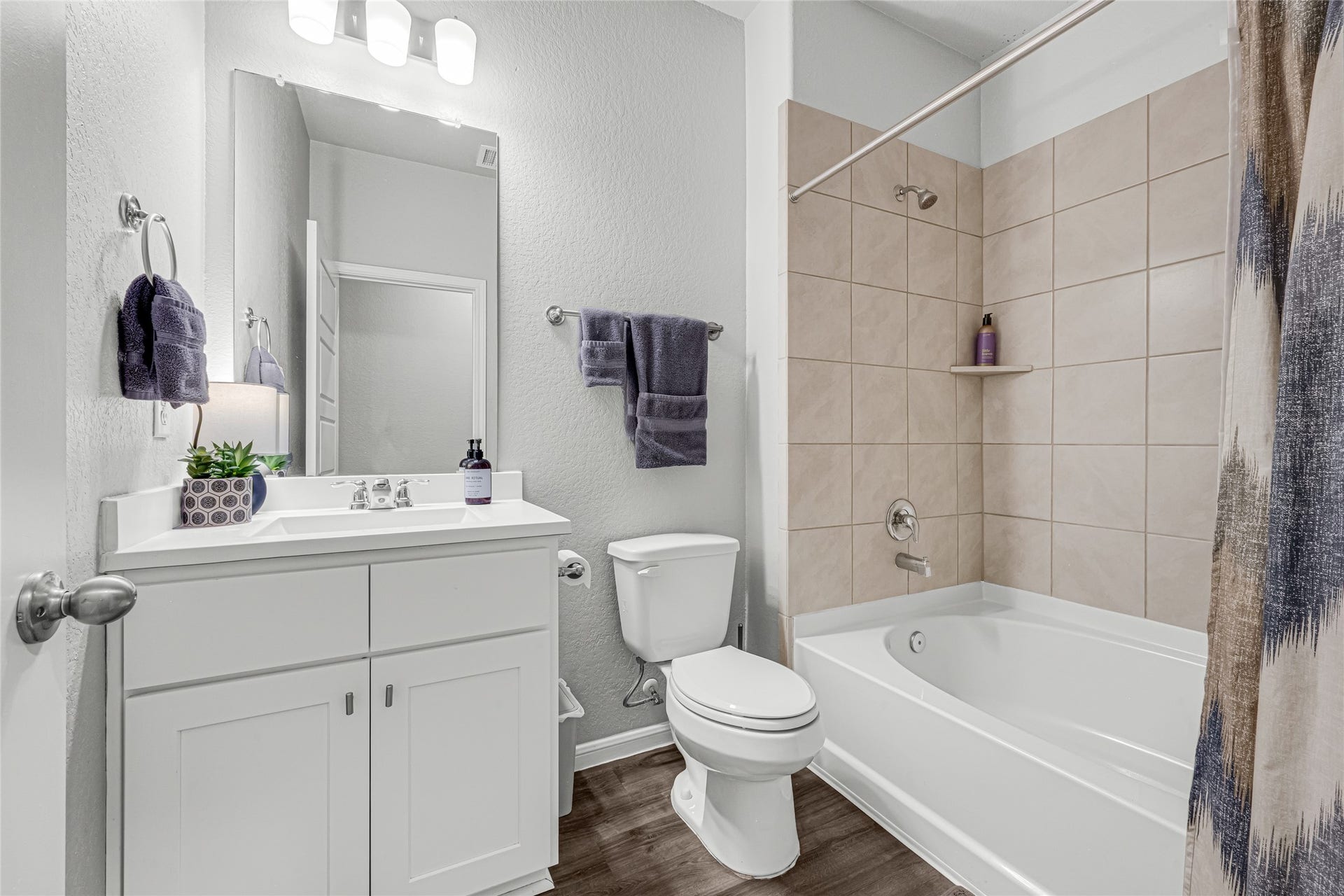 515 Emerald Thicket Lane, Houston, TX 77336 This bathroom features a modern design with a white vanity, large mirror, and sleek lighting with the vinyl plank flooring carrying through. It includes a bathtub with beige tile surround, an toilet, and stylish fixtures, creating a clean and inviting space.