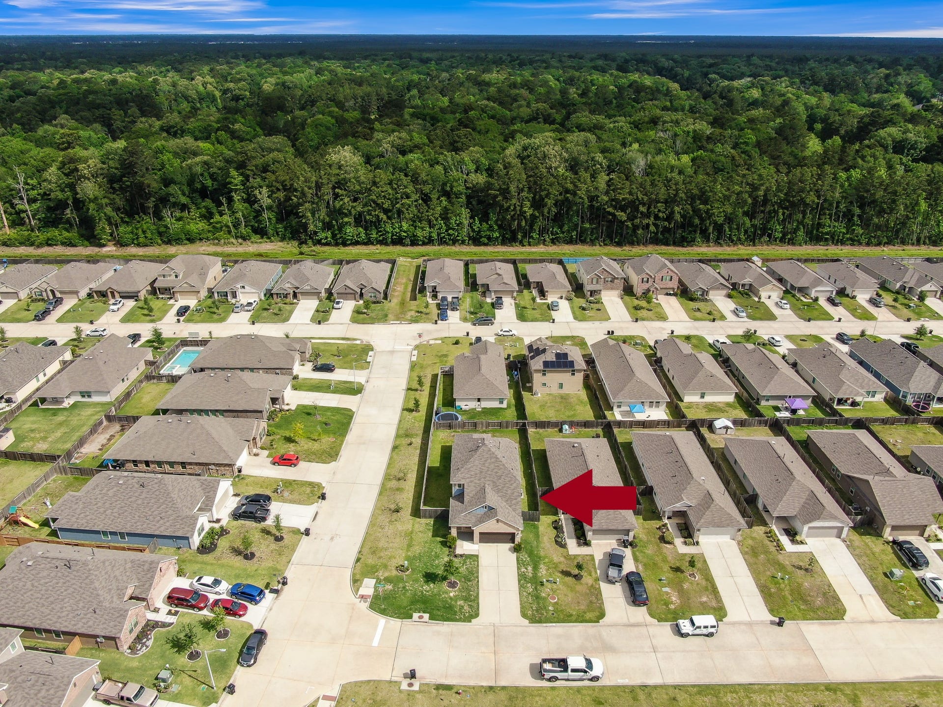22203 Cardinal Crest Lane, New Caney, TX 77357 This aerial photo showcases a suburban neighborhood with rows of single-family homes, each with driveways and small front yards. The area is surrounded by lush greenery, providing a sense of tranquility. An arrow points to a specific house, indicating its location within the community.