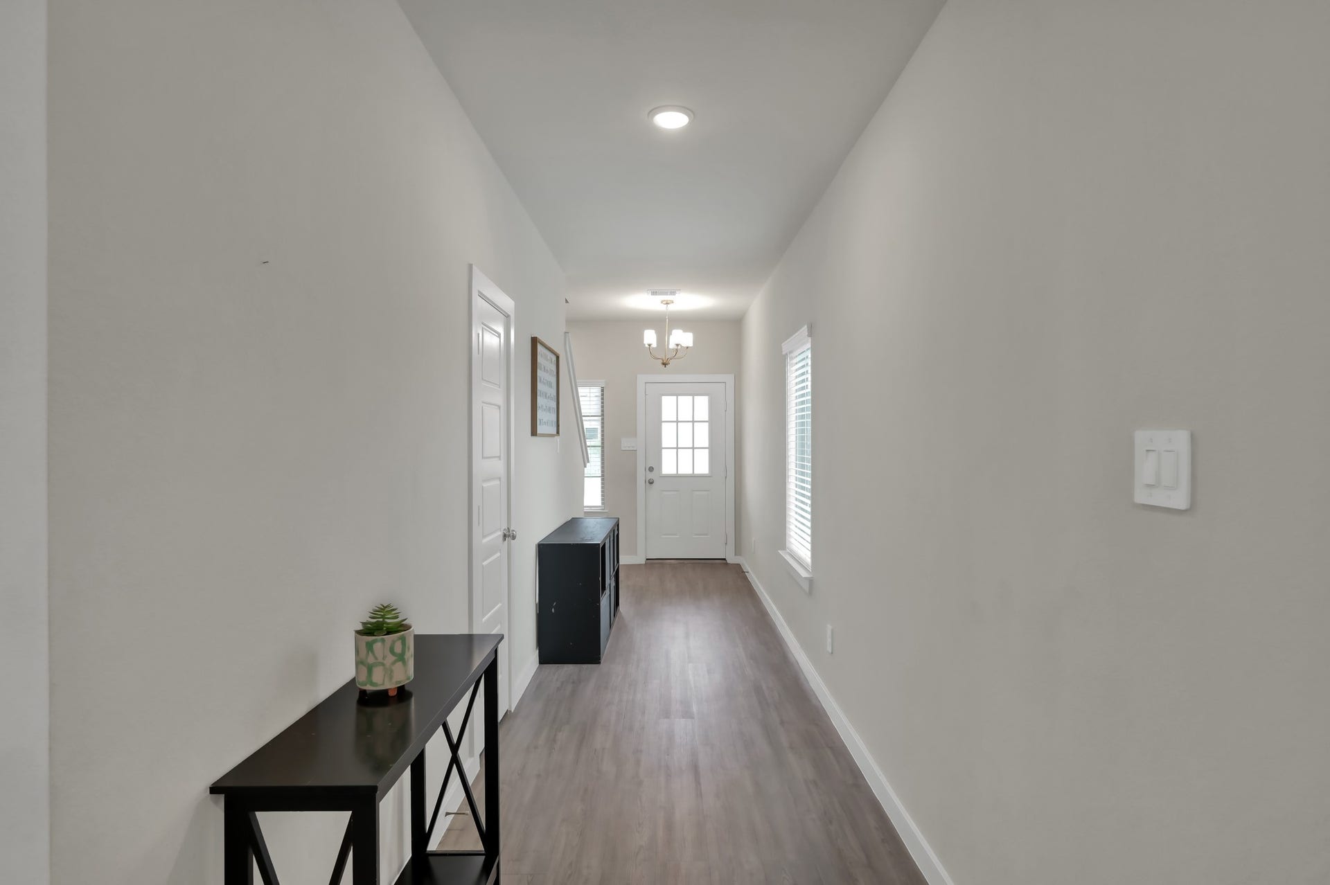 22203 Cardinal Crest Lane, New Caney, TX 77357 Bright and inviting hallway with neutral walls and wood flooring. Features include a modern light fixture, a side table with decor, a window for natural light, and a door leading to other areas of the home.