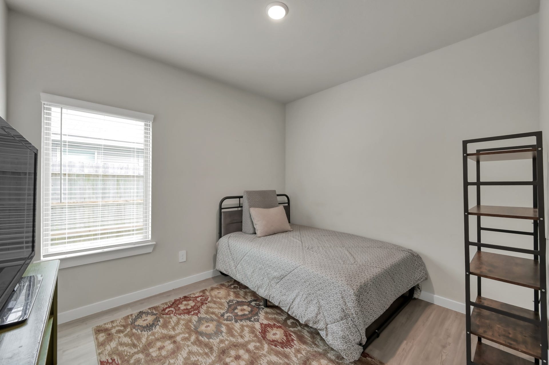 22203 Cardinal Crest Lane, New Caney, TX 77357 This cozy bedroom features a single bed, a window with blinds for natural light, a patterned area rug, and a minimalist metal shelf. The neutral wall and flooring tones create a calm atmosphere.