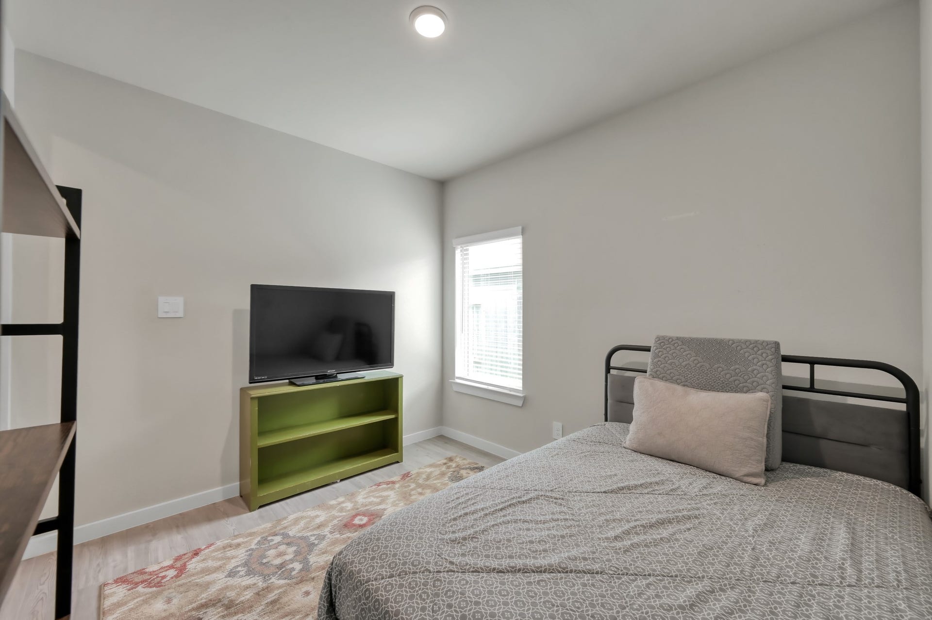 22203 Cardinal Crest Lane, New Caney, TX 77357 This is a cozy, well-lit bedroom featuring a single bed, a TV on a small green stand, and a window with blinds. The neutral decor and simple design make it inviting and functional.