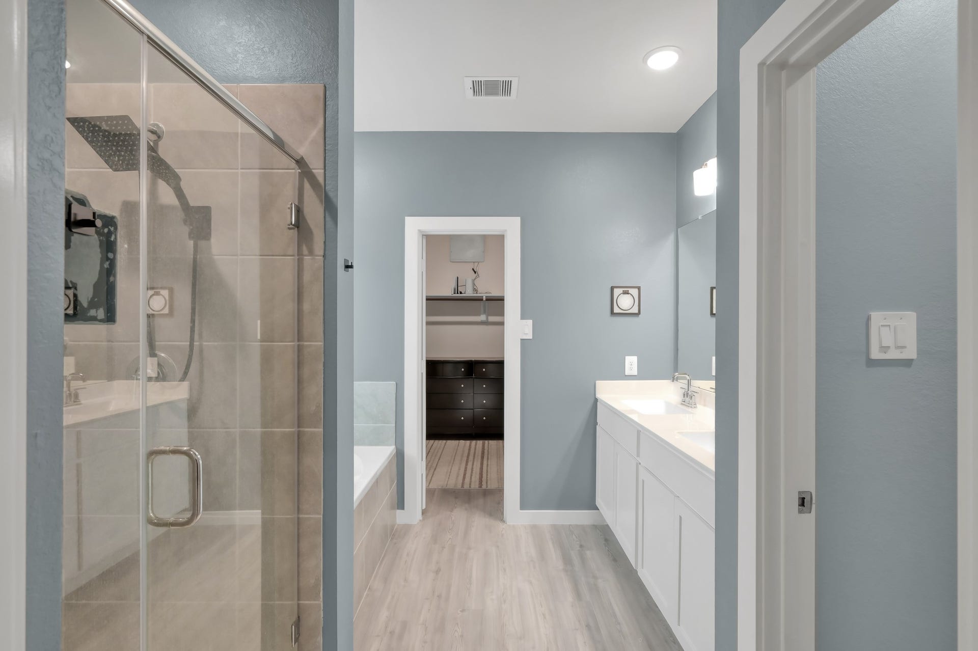 22203 Cardinal Crest Lane, New Caney, TX 77357 This modern bathroom features a glass-enclosed shower, a separate bathtub, dual sinks with a large mirror, soft blue walls, and light wood flooring, creating a bright and spacious feel.