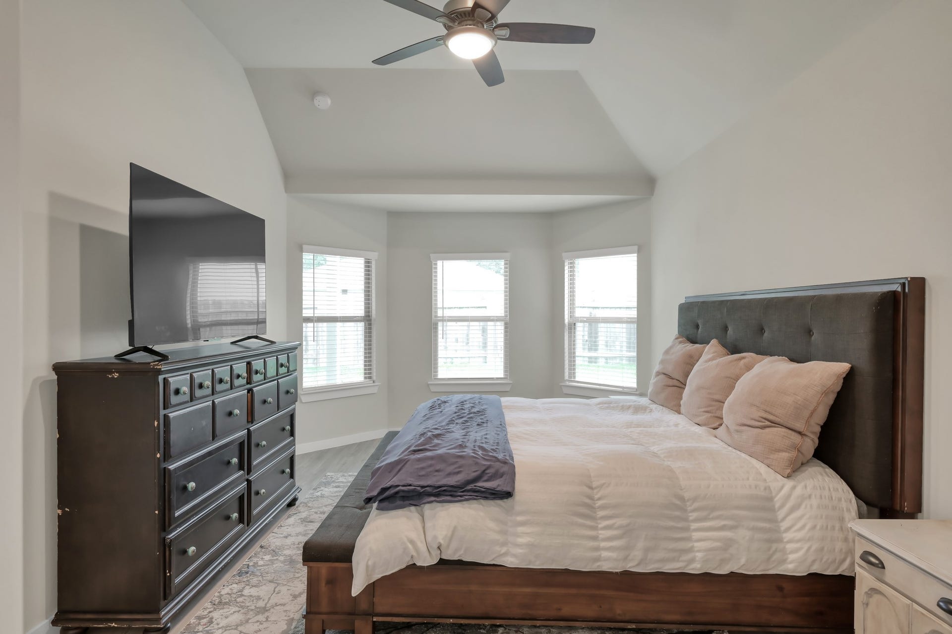 22203 Cardinal Crest Lane, New Caney, TX 77357 This bedroom is spacious and bright, featuring large windows, a high ceiling with a ceiling fan, and a modern design. It includes a comfortable bed and a large dresser with a mounted TV, offering a cozy yet elegant atmosphere.