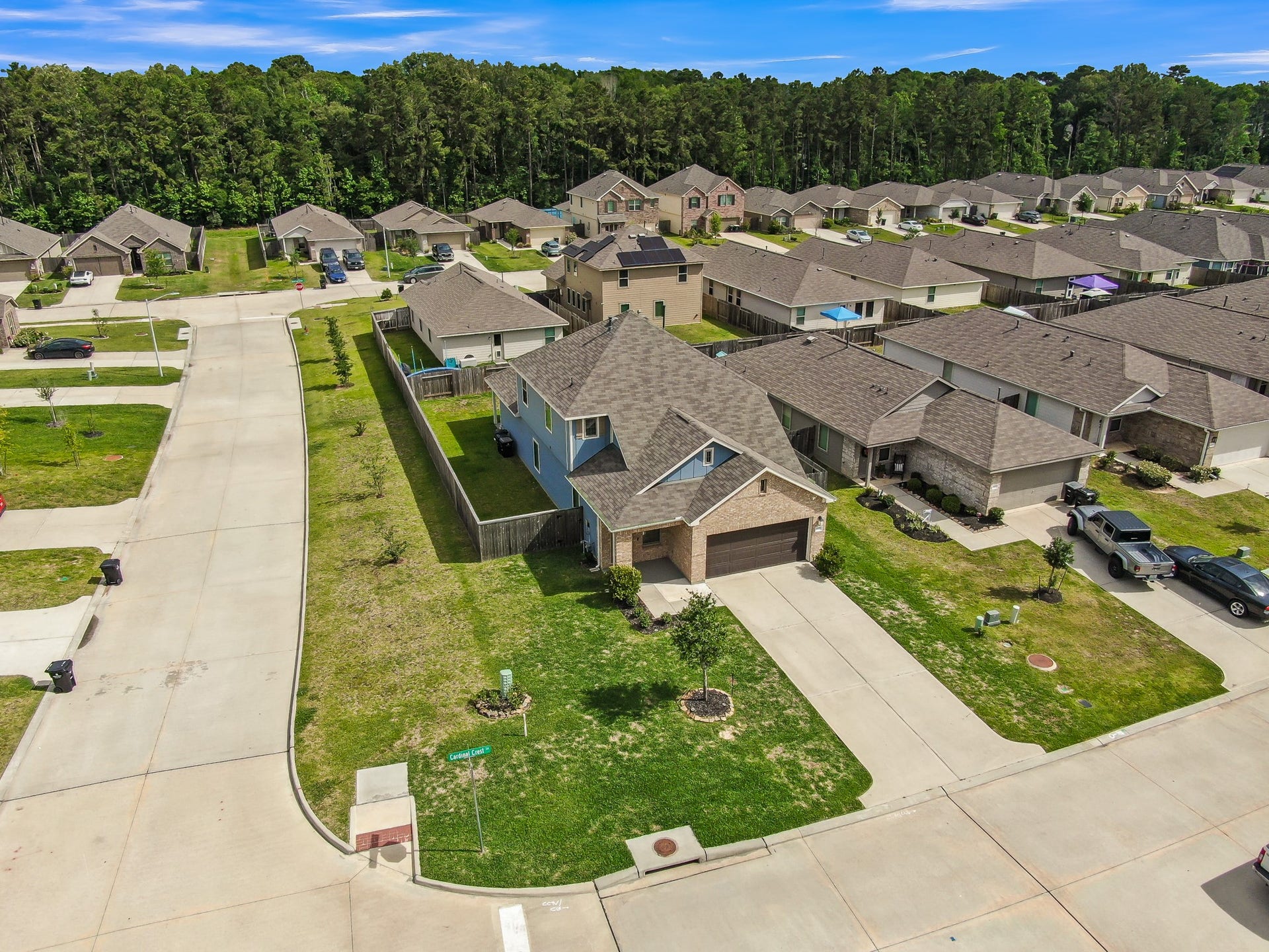 22203 Cardinal Crest Lane, New Caney, TX 77357 Aerial view of a suburban neighborhood featuring a single-story home with a two-car garage and well-maintained lawn. The house is situated on a corner lot with a spacious backyard and privacy fence. The street is lined with similar houses and bordered by a wooded area.