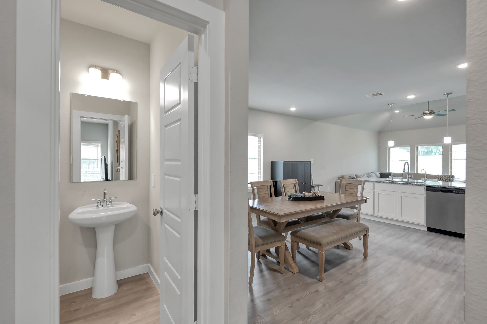 22203 Cardinal Crest Lane, New Caney, TX 77357 An open-concept living space featuring a dining area with a wooden table set and a modern kitchen with white cabinetry and stainless steel appliances. There's also a glimpse of a small bathroom with a pedestal sink and mirror. The area has light wood flooring and ample natural light from large windows.