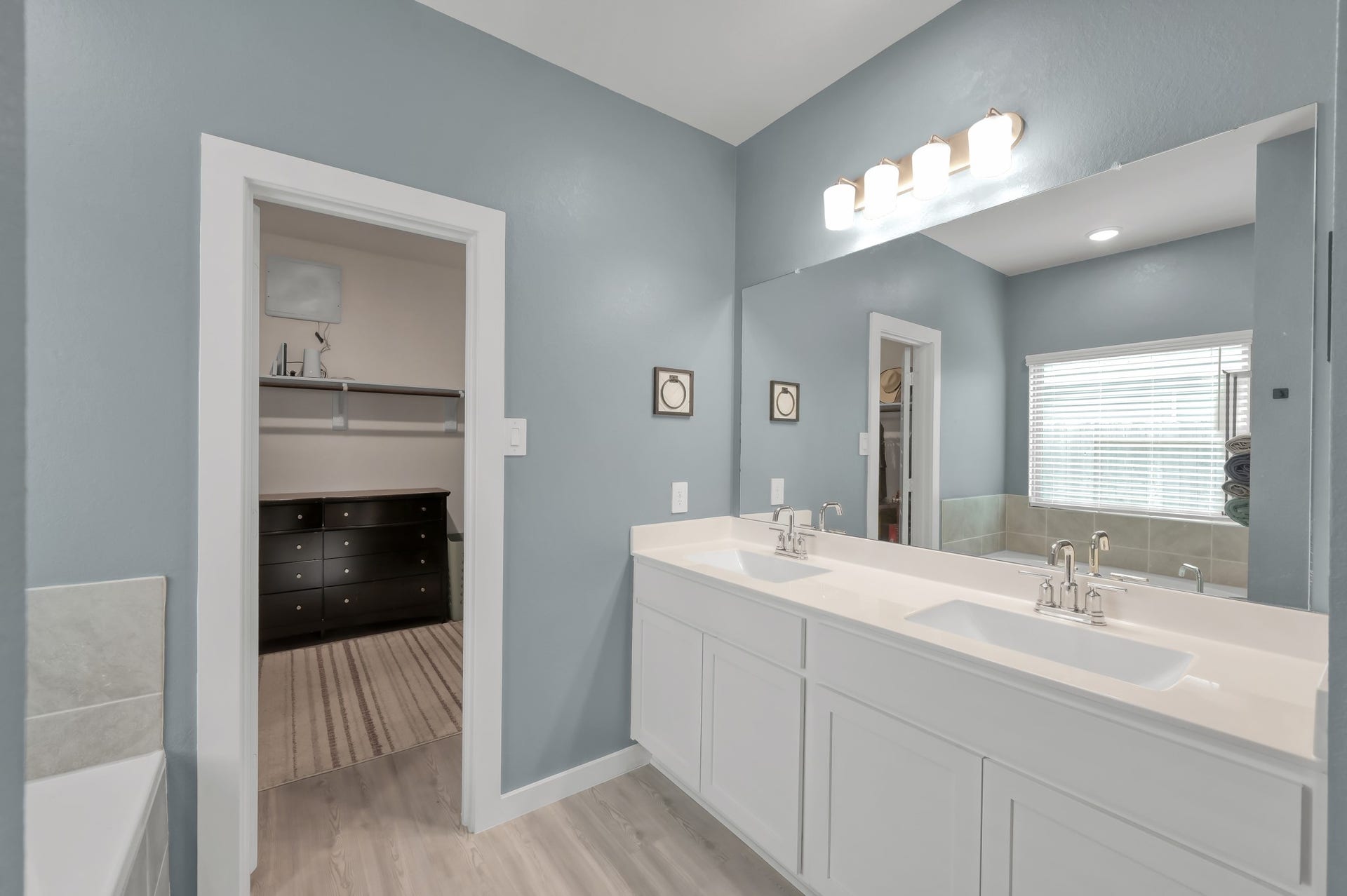 22203 Cardinal Crest Lane, New Caney, TX 77357 A modern bathroom with a dual-sink vanity, sleek white cabinets, and elegant fixtures. The room features a large mirror, soft blue walls, and ample natural light from a window. A walk-in closet is visible through an open door, offering additional storage space.