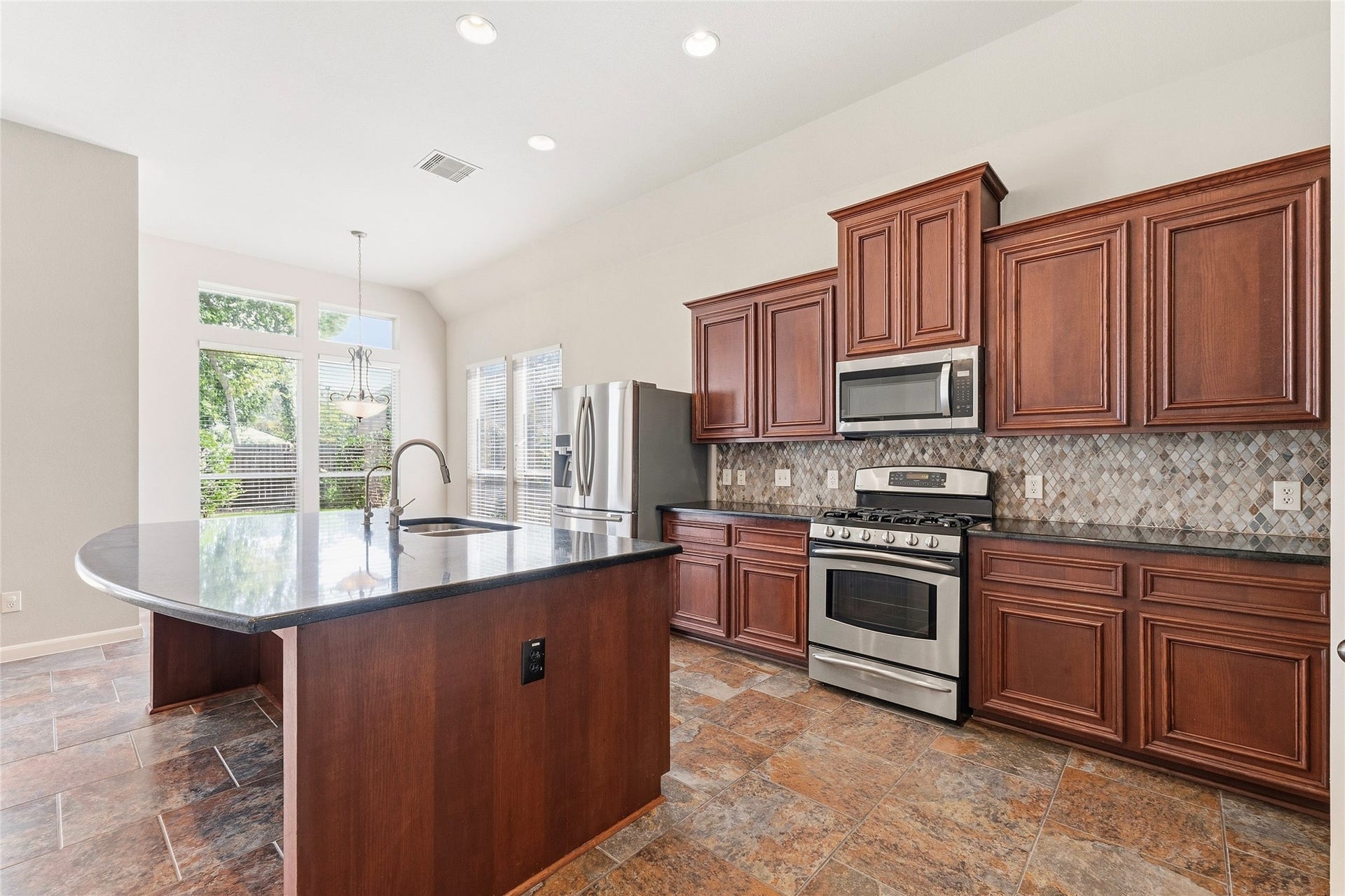Spacious kitchen with rich wooden cabinetry, sleek granite countertops, and modern stainless steel appliances. The large island offers ample workspace, while abundant natural light pours in through the windows, enhancing the warm, inviting atmosphere.