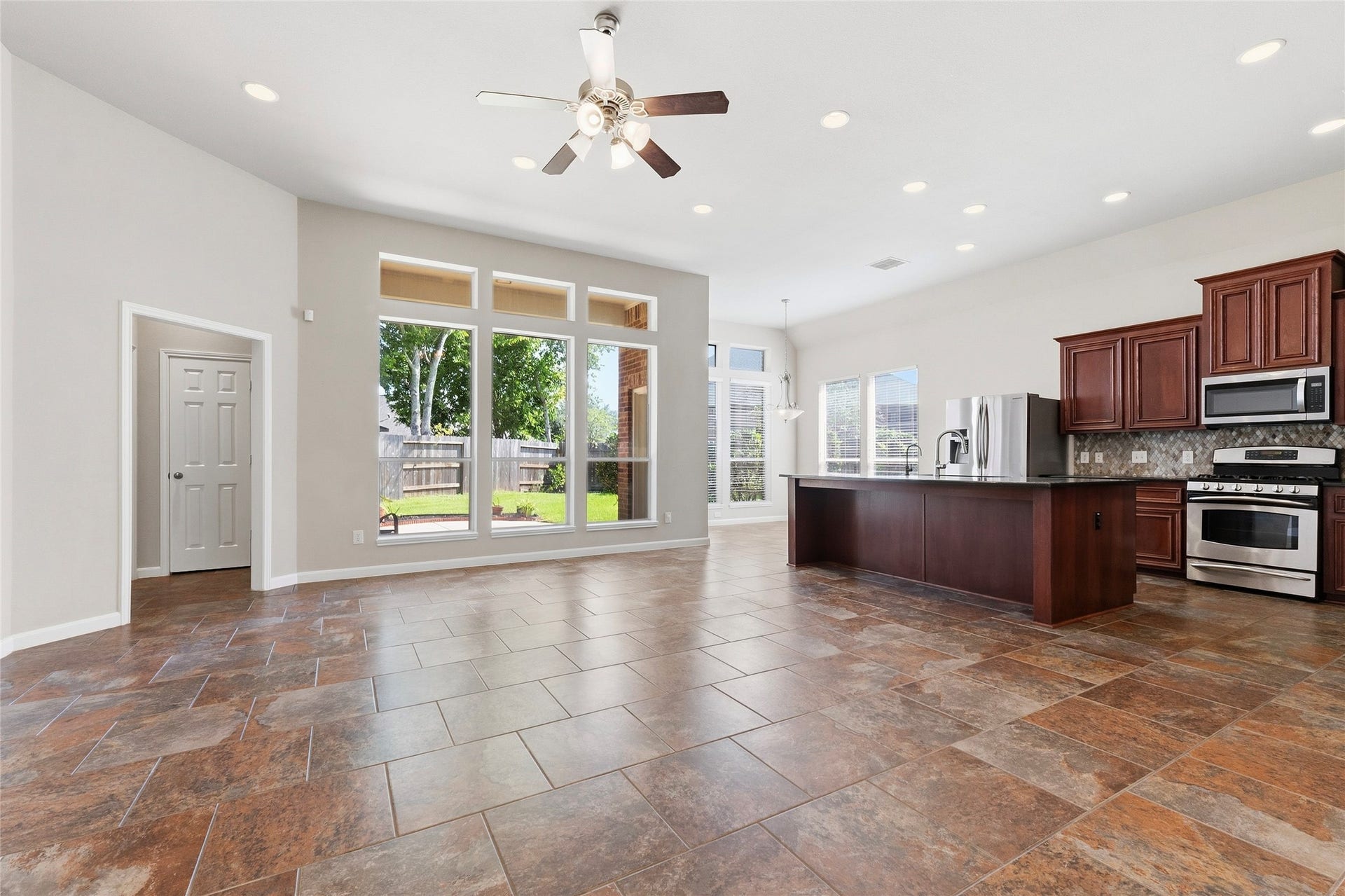 Spacious open-concept living area with a modern kitchen, featuring stainless steel appliances and rich wood cabinetry, complemented by large windows that bring in abundant natural light and overlook the backyard.