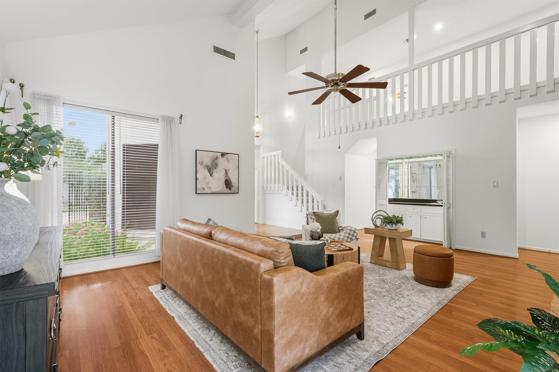 12219 Ravenmoor Drive, Houston, TX 77077 Spacious living room with high ceilings, featuring a cozy leather sofa, elegant hardwood floors, and abundant natural light from large windows. A stylish staircase and modern ceiling fan add to the contemporary charm.