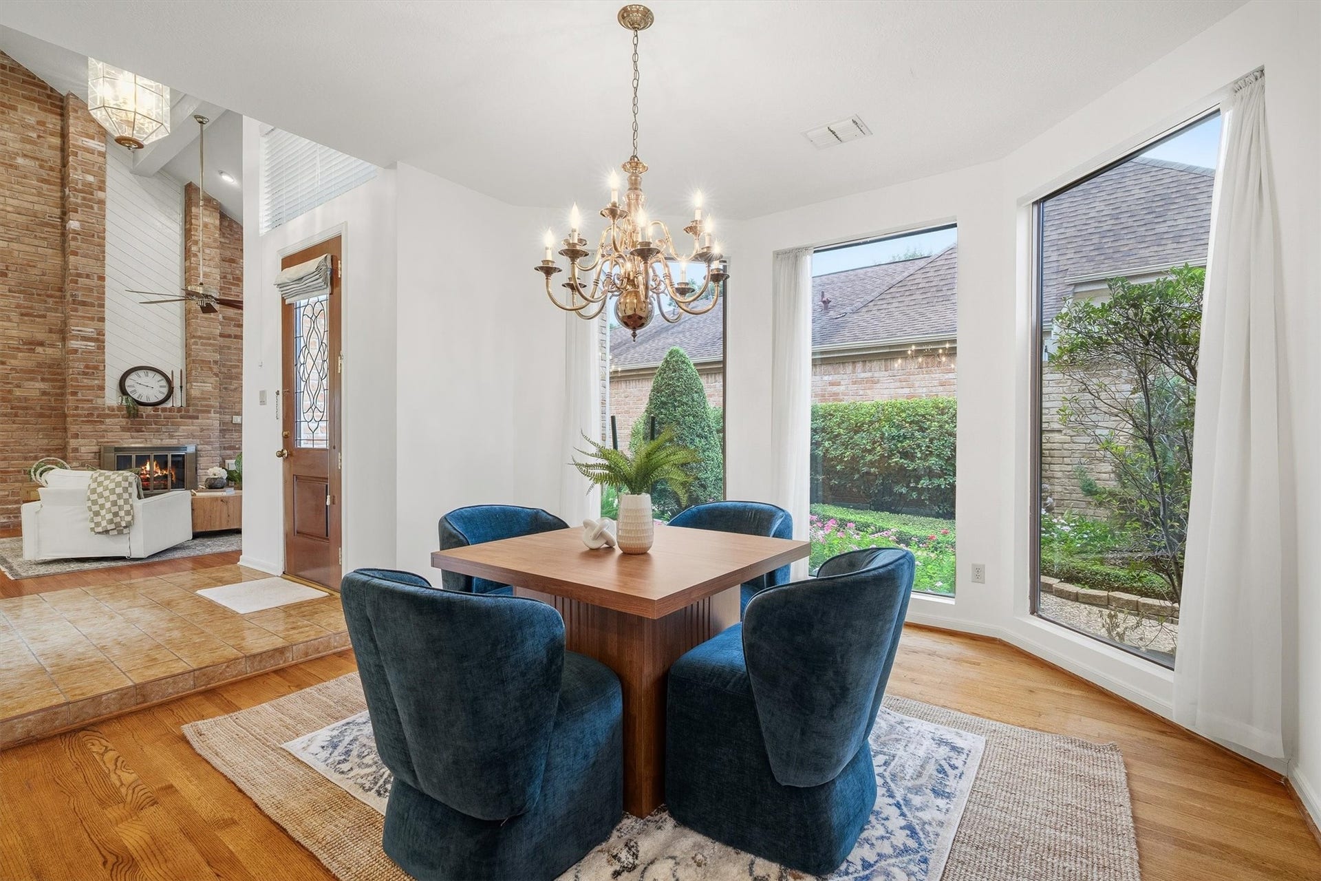 12219 Ravenmoor Drive, Houston, TX 77077 Great dining area with large windows providing garden views, complemented by a charming chandelier and plush seating. Adjacent is a cozy living space featuring a brick fireplace and high ceilings for an inviting atmosphere.