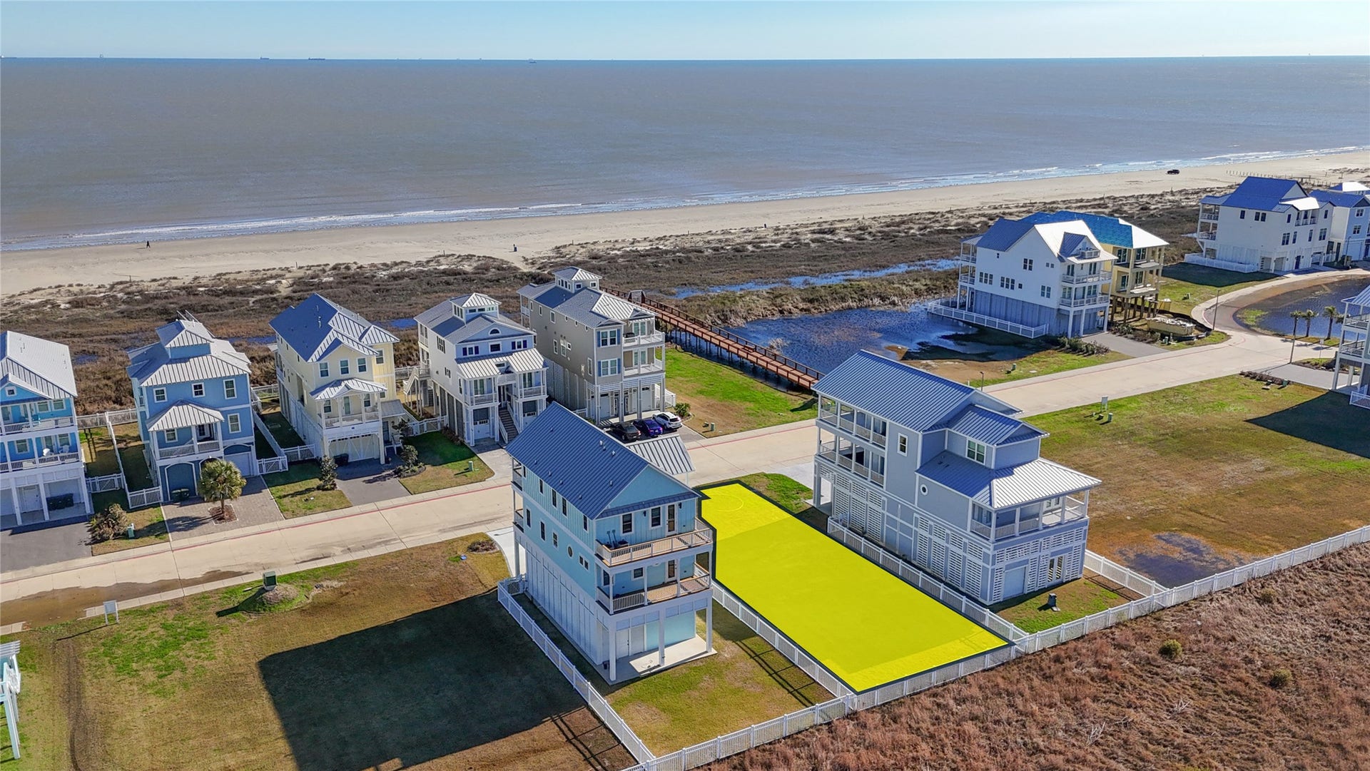 Looking across the community toward the Gulf, this aerial view showcases the open setting surrounding the homesite and the impressive coastal residences already established nearby. The cleared building site offers a blank canvas for a thoughtfully designed custom home, with a 50-foot frontage and approximately 120 feet of depth providing flexibility for an elevated residence, outdoor living areas and coastal-oriented architecture.