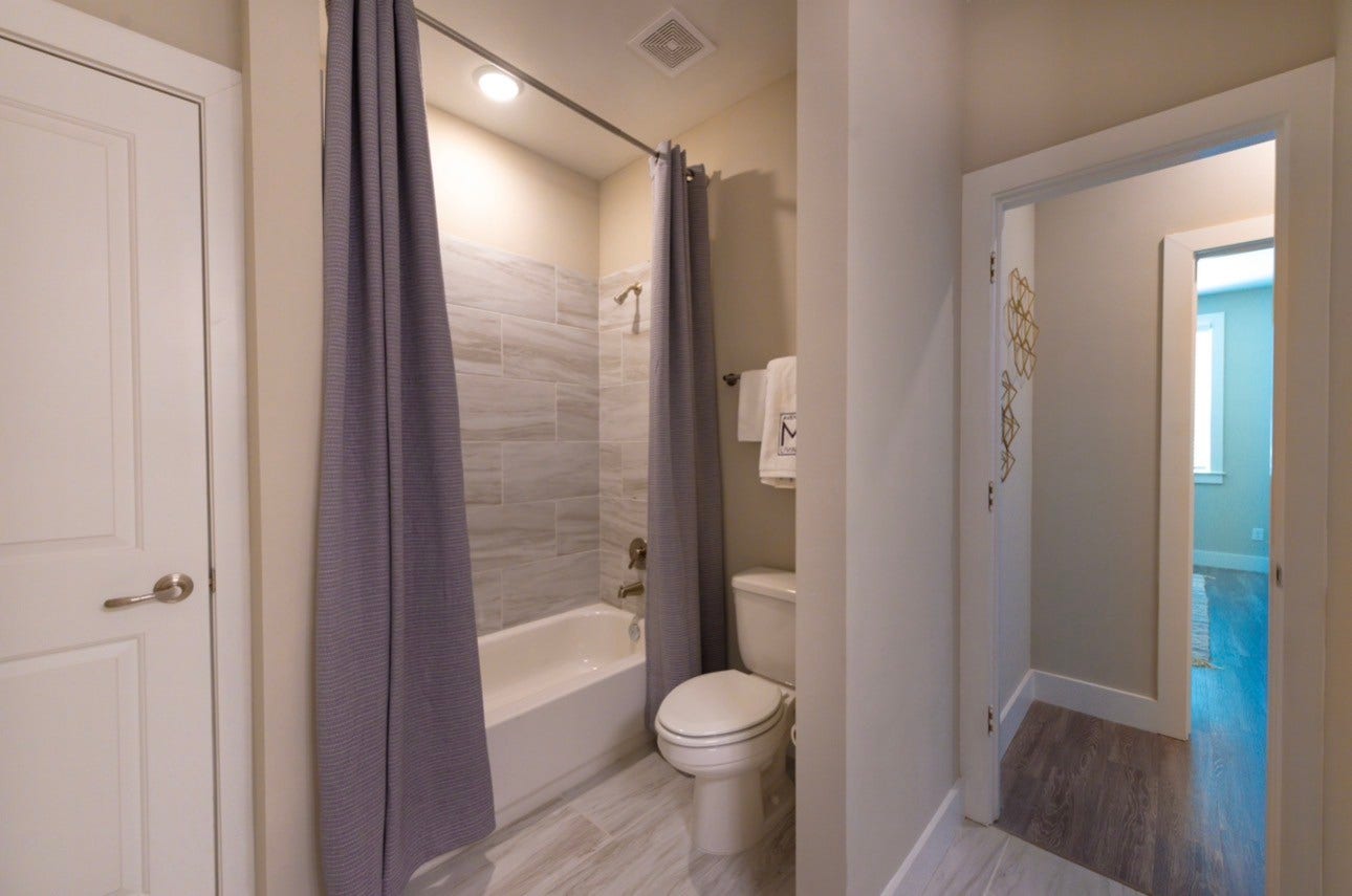 Charming bathroom featuring a modern tub with a gray tile surround, complemented by sleek flooring and a convenient hallway connection to the bedroom