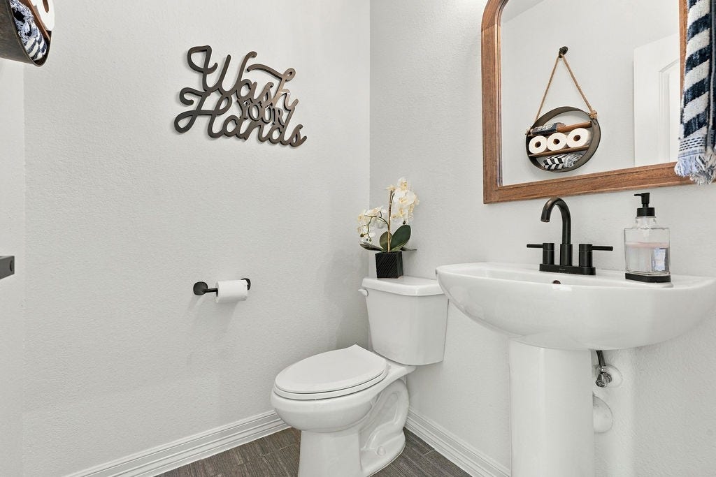 9918 Hubble Drive, Iowa Colony, TX 77583 Charming and compact half bathroom featuring a sleek pedestal sink, modern faucet, and decorative mirror. Stylish wall art and convenient storage add personality and practicality to the space.