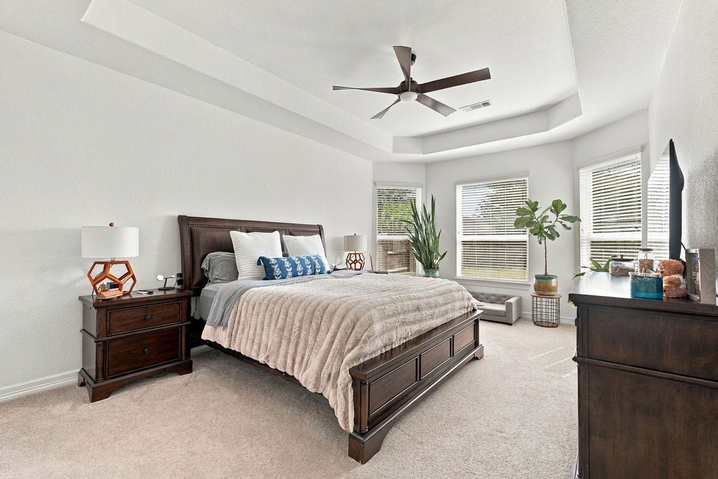 9918 Hubble Drive, Iowa Colony, TX 77583 Spacious primary bedroom with a tray ceiling, large windows for natural light, and elegant decor featuring a cozy bed, stylish nightstands, and lush plants.