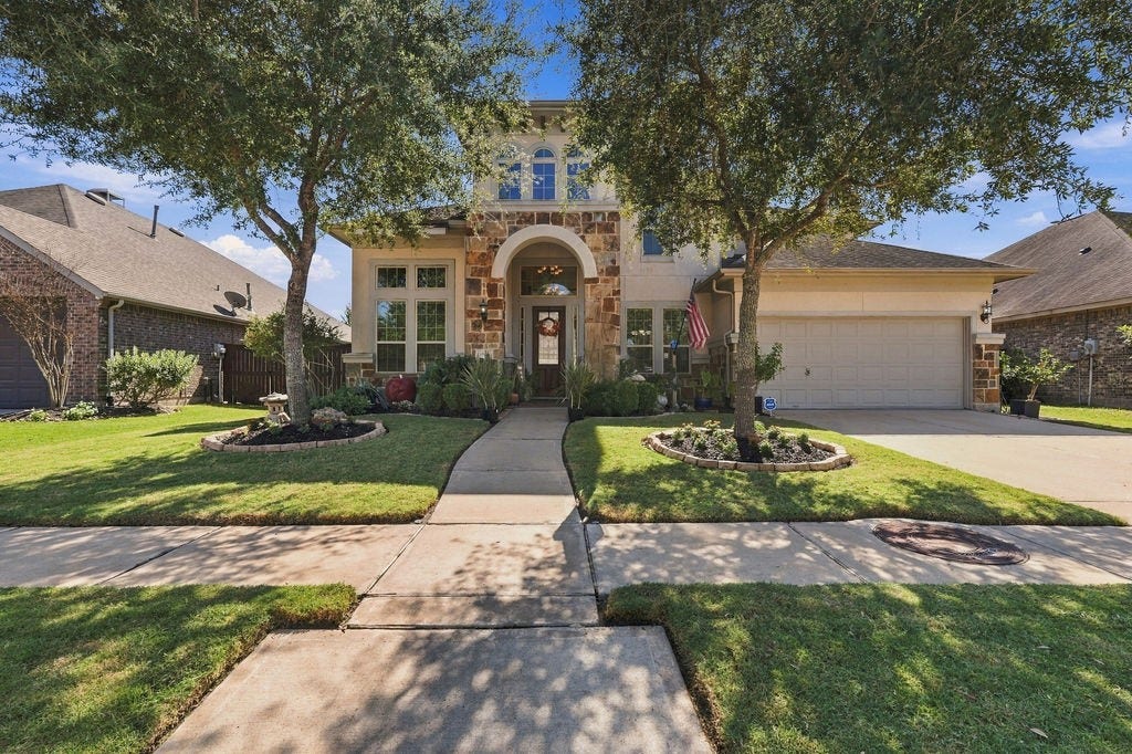 9918 Hubble Drive, Iowa Colony, TX 77583 Stunning two-story home with a stone-accented facade, framed by mature trees and manicured landscaping, featuring a spacious driveway and welcoming entryway.