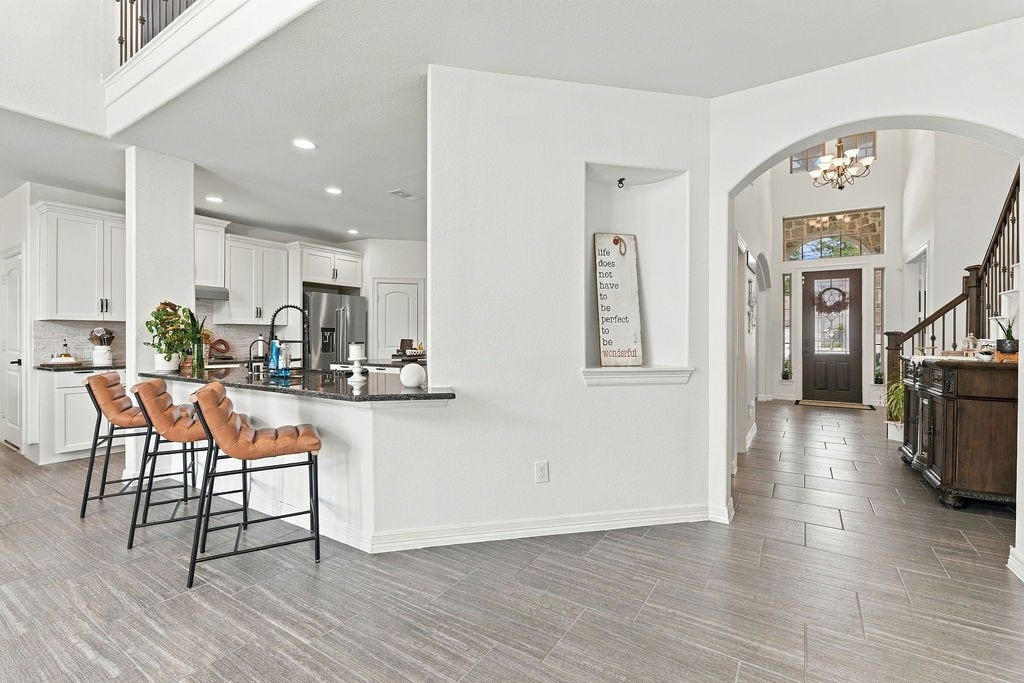 9918 Hubble Drive, Iowa Colony, TX 77583 A modern open-concept kitchen and entryway featuring sleek white cabinetry, a stylish breakfast bar with room for multiple barstools, and elegant tile flooring leading to a welcoming front door.