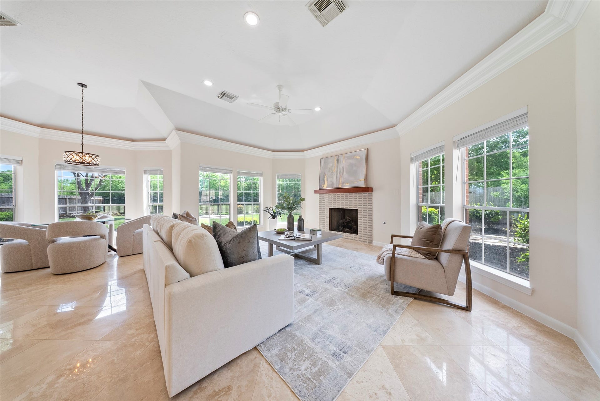 Bright and inviting living room featuring a tray ceiling, fireplace focal point, and walls of windows overlooking the backyard & nature backdrop.