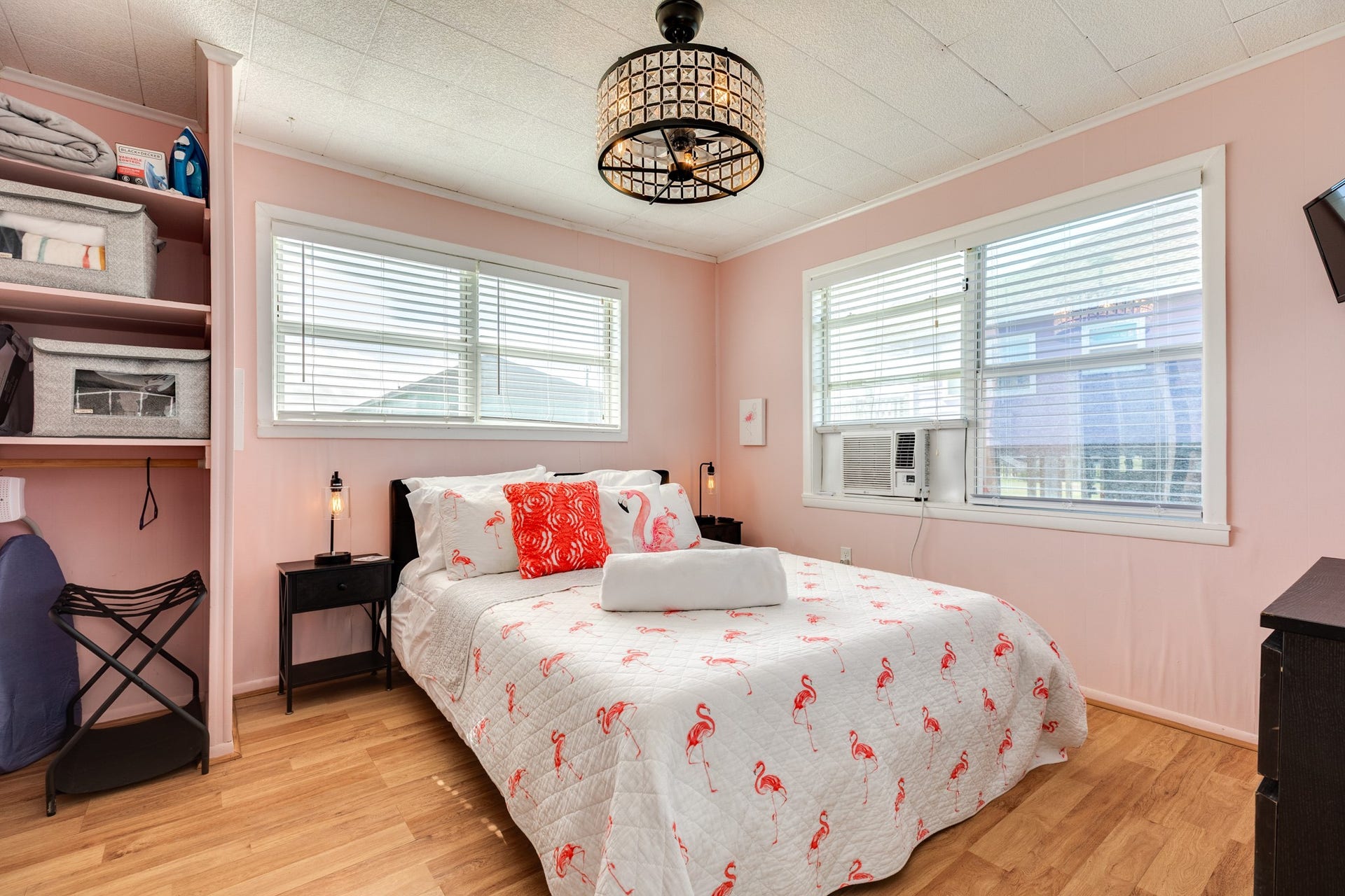 Cheerful bedroom offers pink paneled walls, windows on two sides, wood-look flooring, open closet storage, and a decorative ceiling fixture.