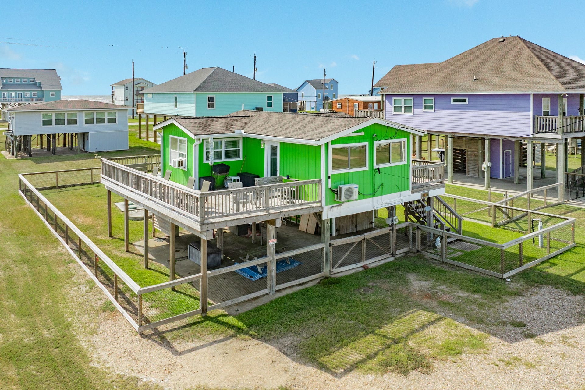 Angled aerial view highlights the generous deck, elevated construction, exterior stair access, and fenced grounds surrounding the property.