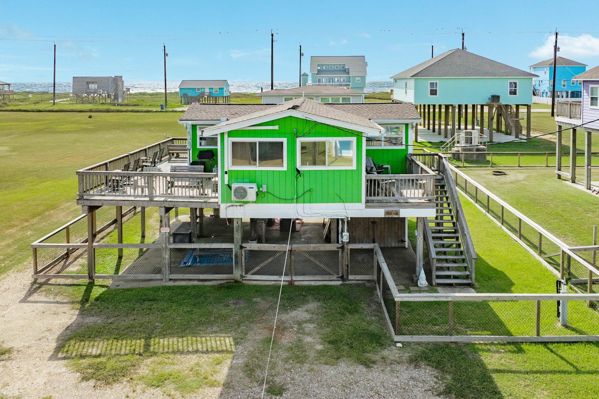 Elevated coastal home features vibrant green siding, expansive wraparound decking, and a large fenced yard with covered space beneath the home.