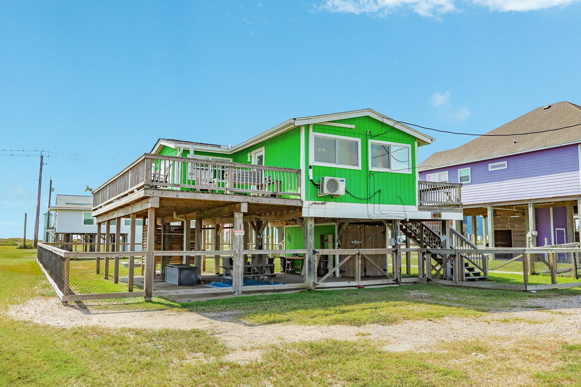 Ground-level view showcases the elevated coastal home, expansive wood decking, fenced yard, and covered space beneath the residence.