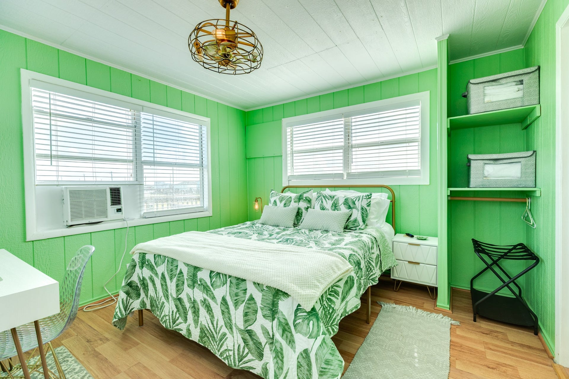 Bright bedroom features green paneled walls, windows on two sides, wood-look flooring, open closet storage, and space for a small desk.
